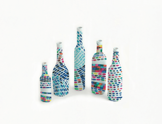 Five ceramic bottles are arranged in a row against a white background. Each bottle is decorated with a mosaic of small, multicolored dots and lines. The bottles vary in height, with the tallest in the center and the shortest on the left. The white caps at the top of each bottle contrast with the colorful patterns.