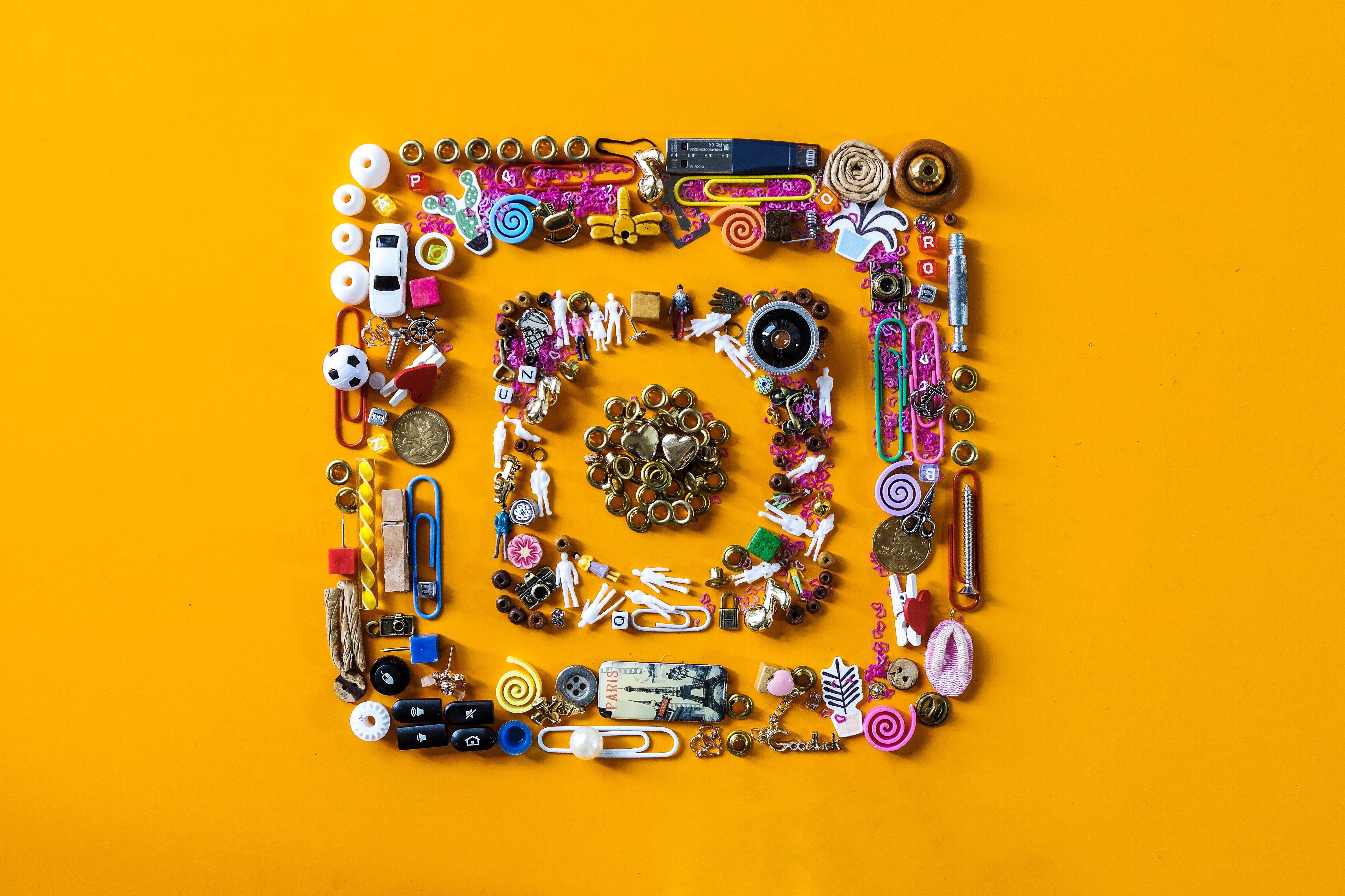 A creative top-down flat lay photo of the Instagram logo made from a colorful collection of miscellaneous small objects on a bright yellow background. The iconic camera shape is formed using items like paper clips, tiny human figurines, beads, buttons, charms, coins, and miniature toys.
