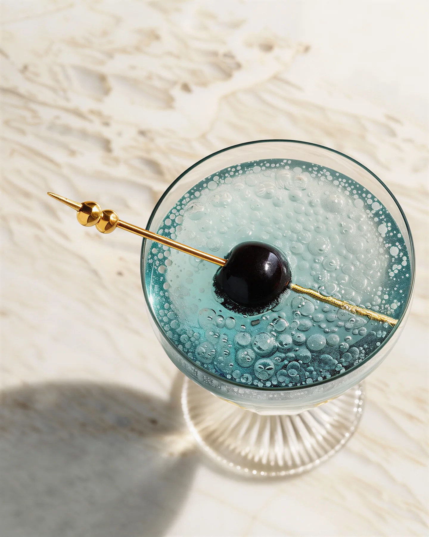 Turquoise cocktail in a coupe with a black cherry on a gold pick