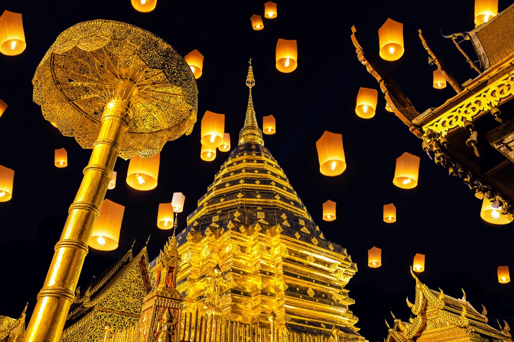 Golden temple structures and intricately designed umbrellas illuminated at night, with numerous floating lanterns ascending into the sky, creating a magical and serene atmosphere at a traditional festival in Thailand.