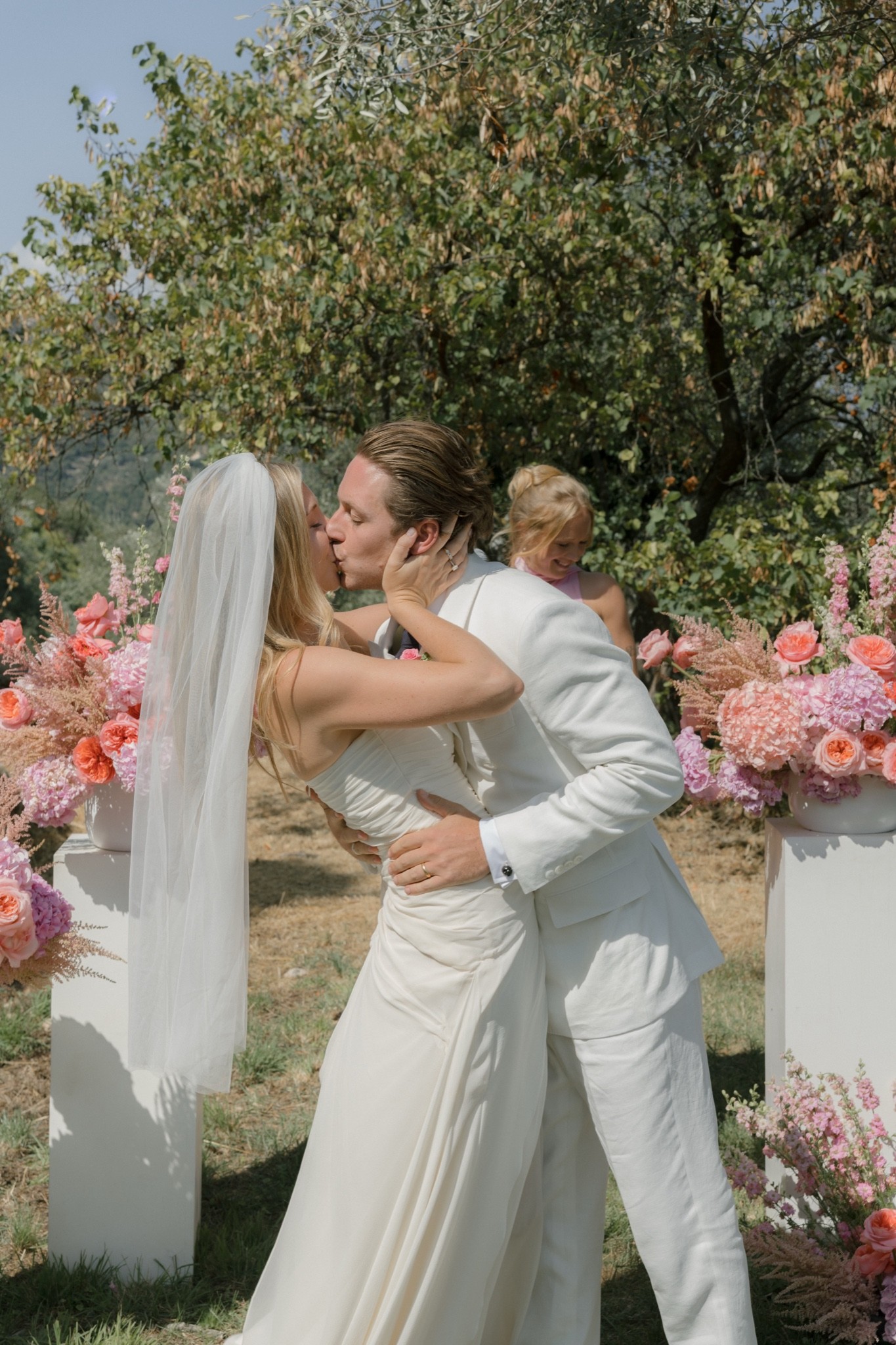 The couple kisses beside pink and orange flower arrangements under an olive tree