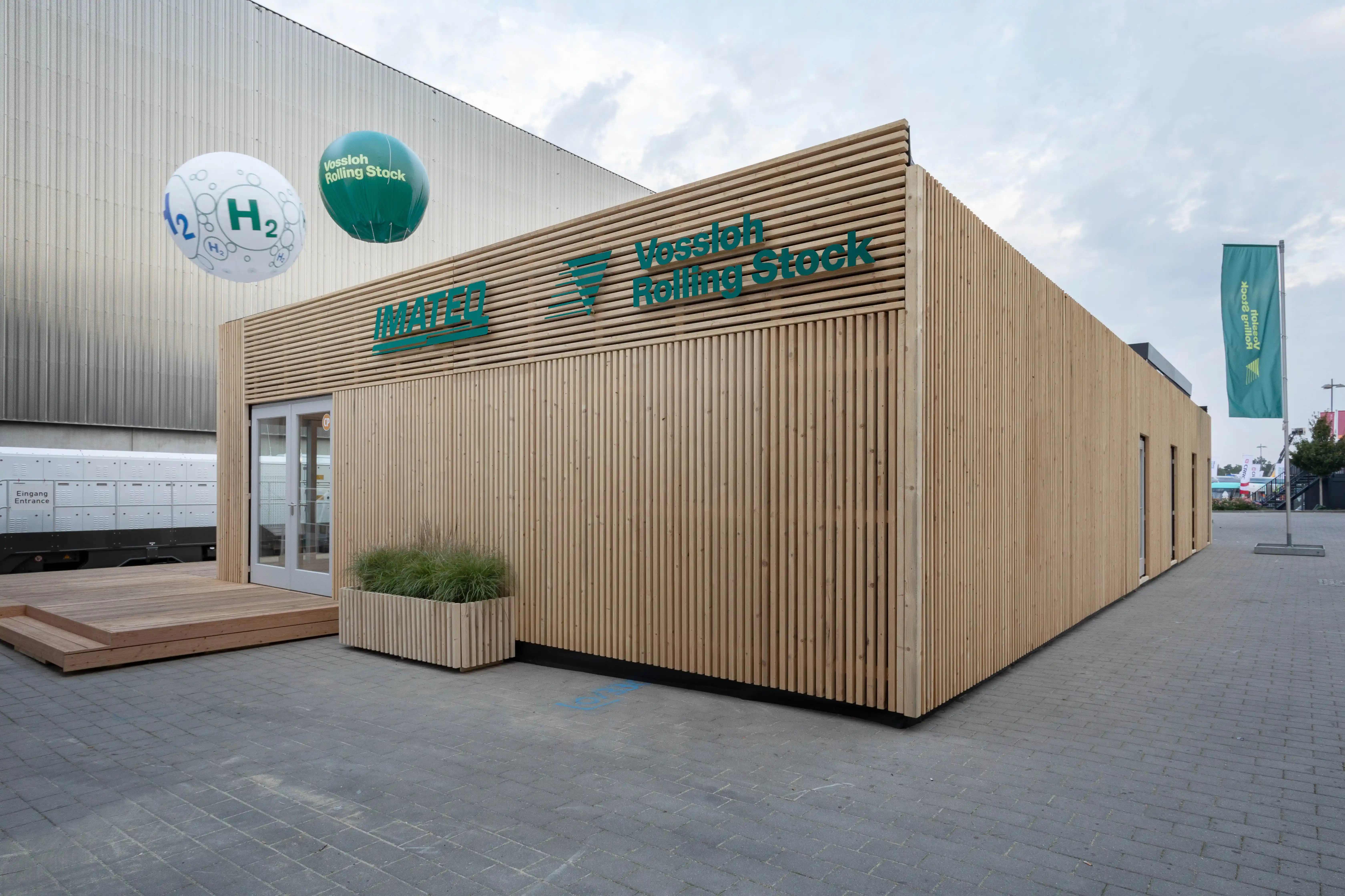 Wooden pavilion of the Vossloh Rolling Stock house at InnoTrans with slatted larch facade, green logos and balloons above.