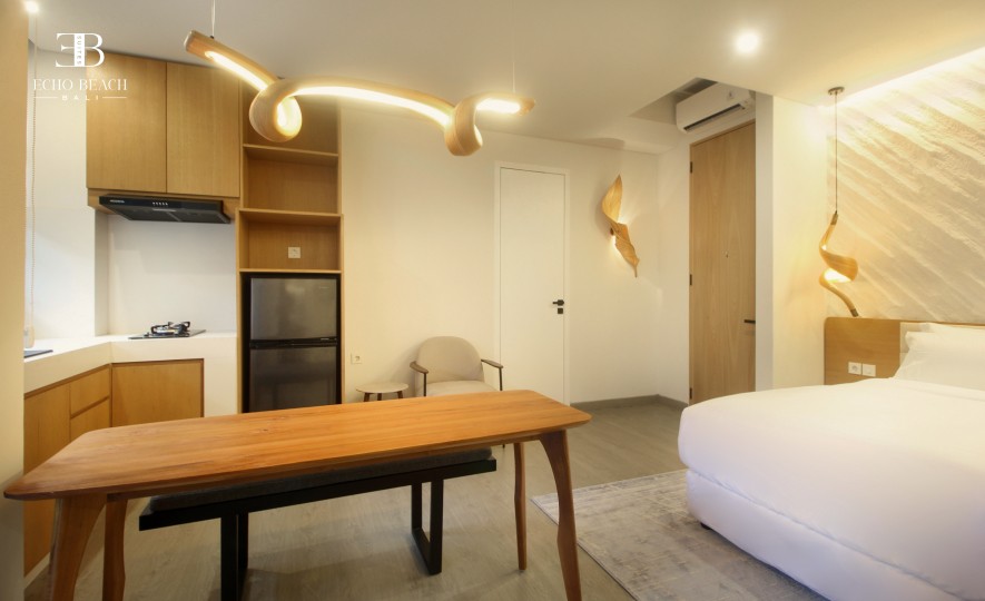 Open-plan kitchenette and dining area featuring a wooden table and bench, a refrigerator, and unique pendant lighting, leading to the bedroom at the Echo Beach Suite in Canggu, Bali.