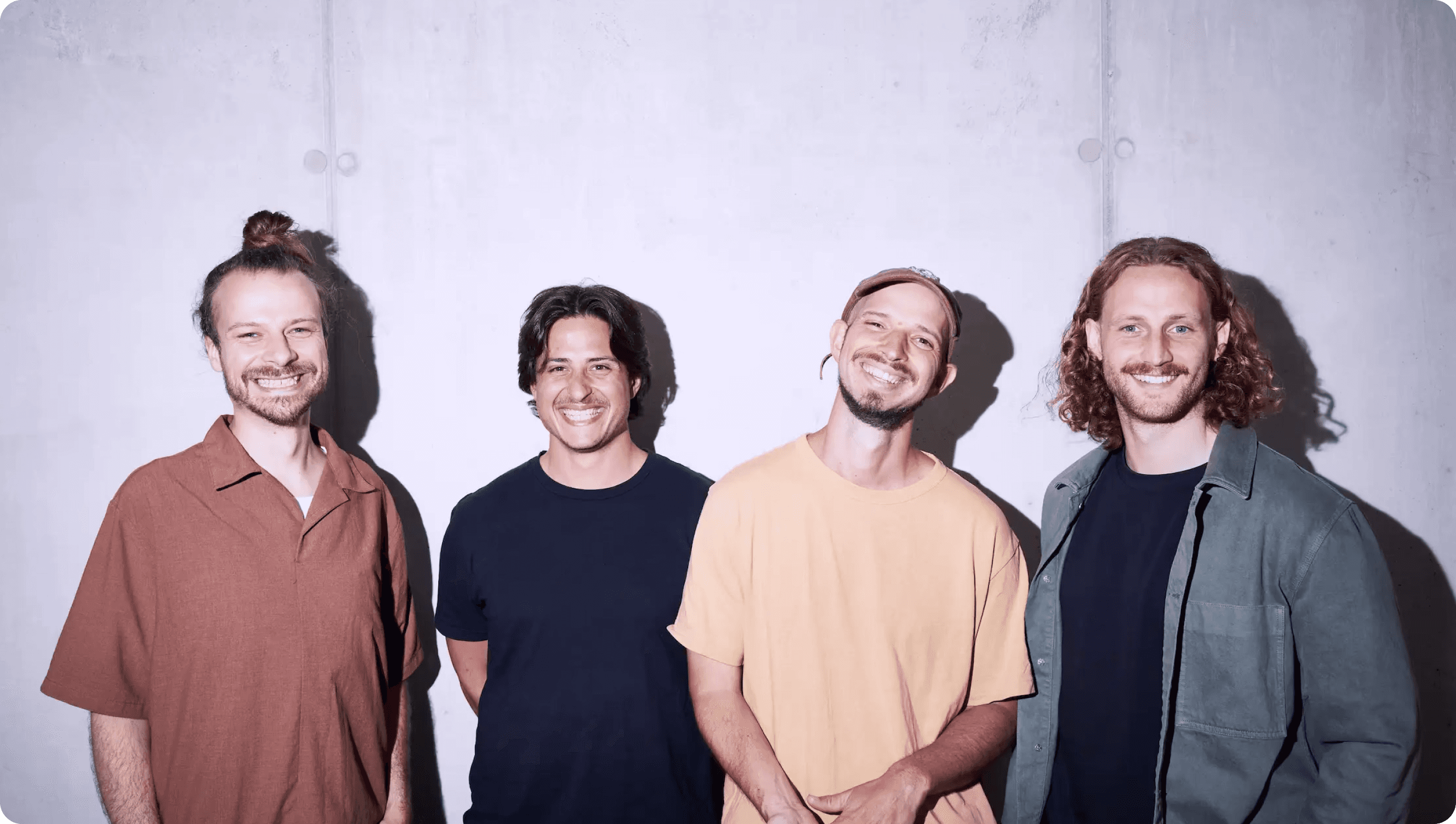 Vier Männer stehen vor einem schlichten Hintergrund und lächeln alle.