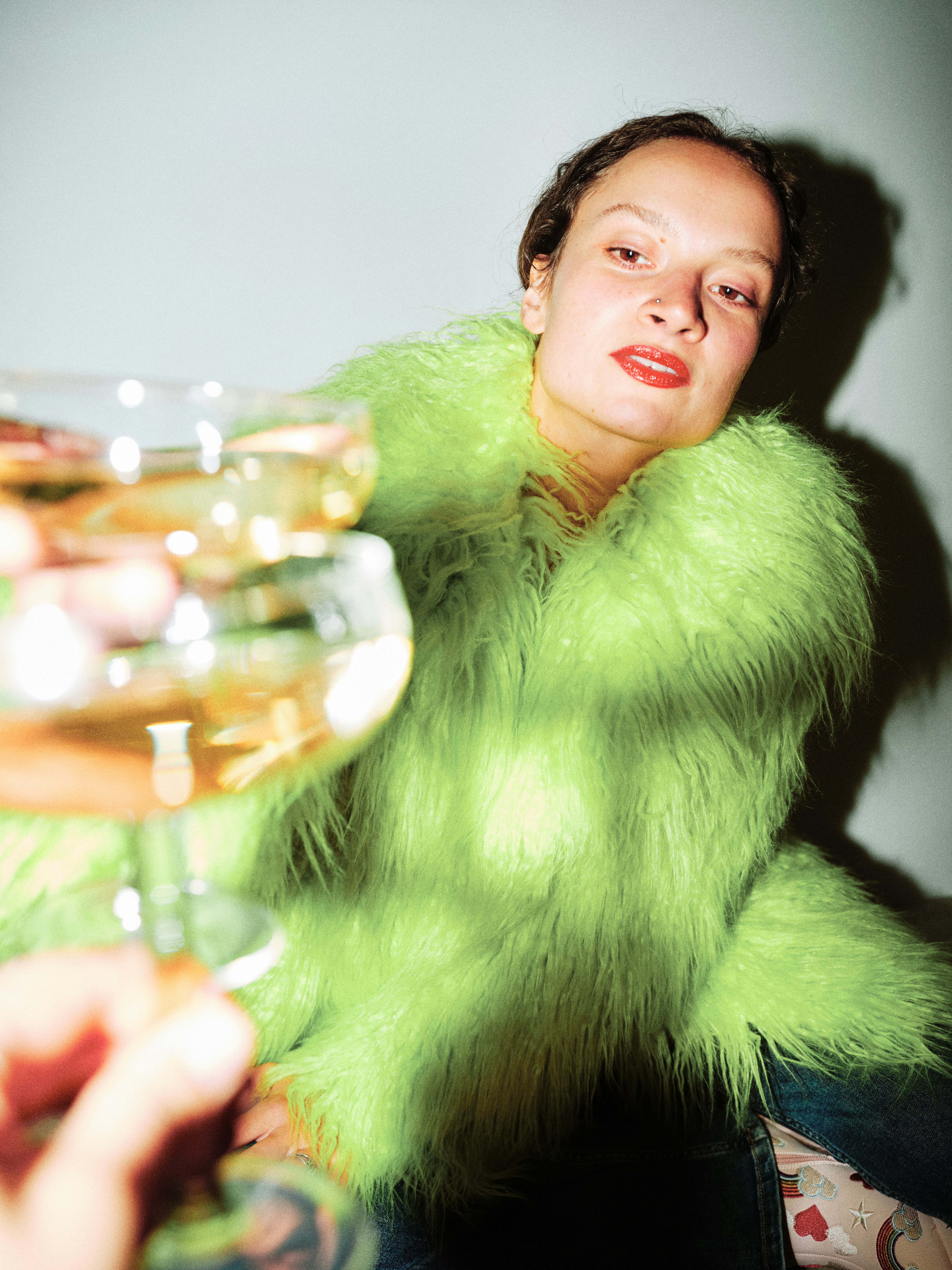 Candid editorial portrait of a model in a lime green faux fur coat with red lipstick, two champagne glasses clinking in the foreground