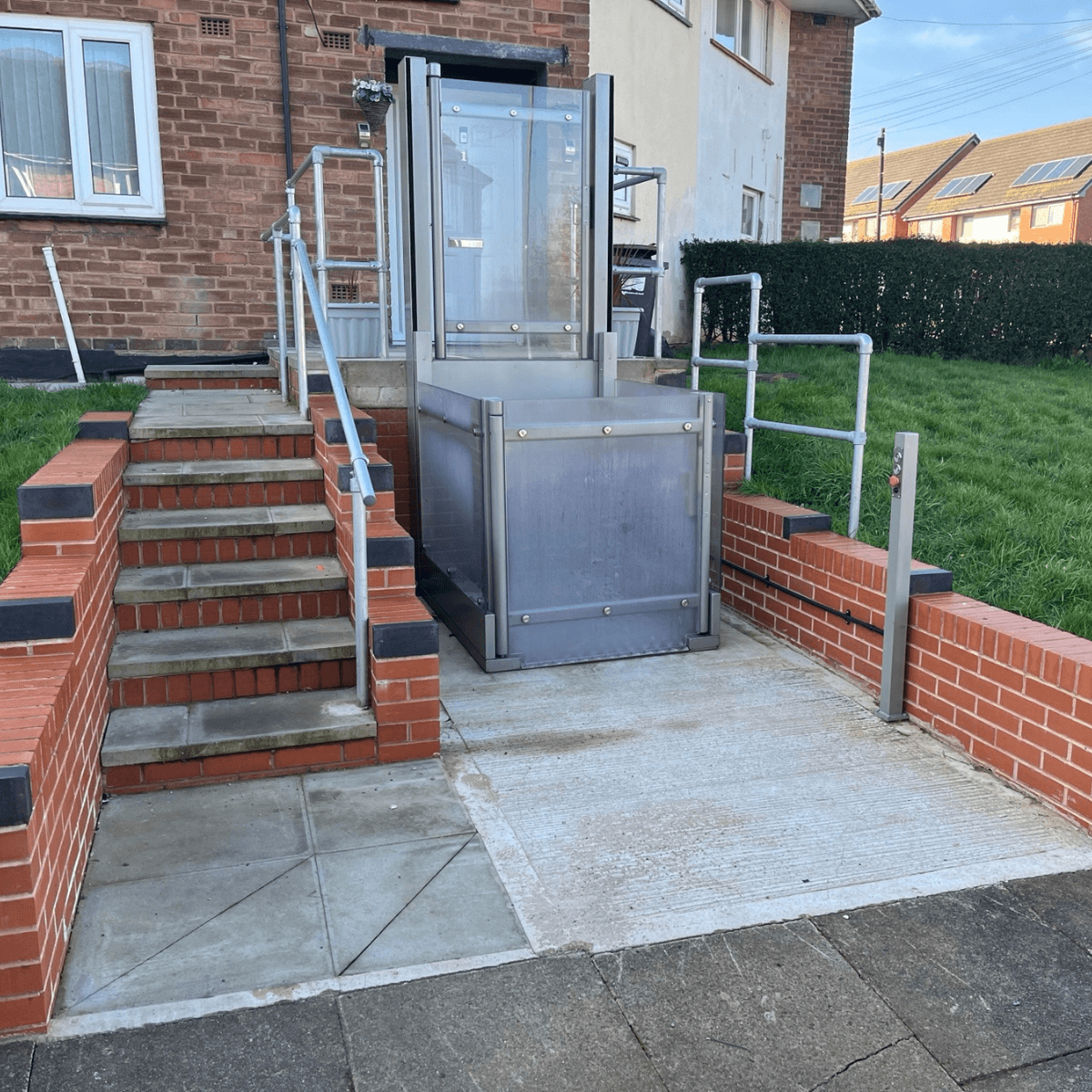 Outdoor vertical platform lift with full glass enclosure installed against a red brick garden wall — stainless steel frame with anti-slip chequer plate platform at lower garden level