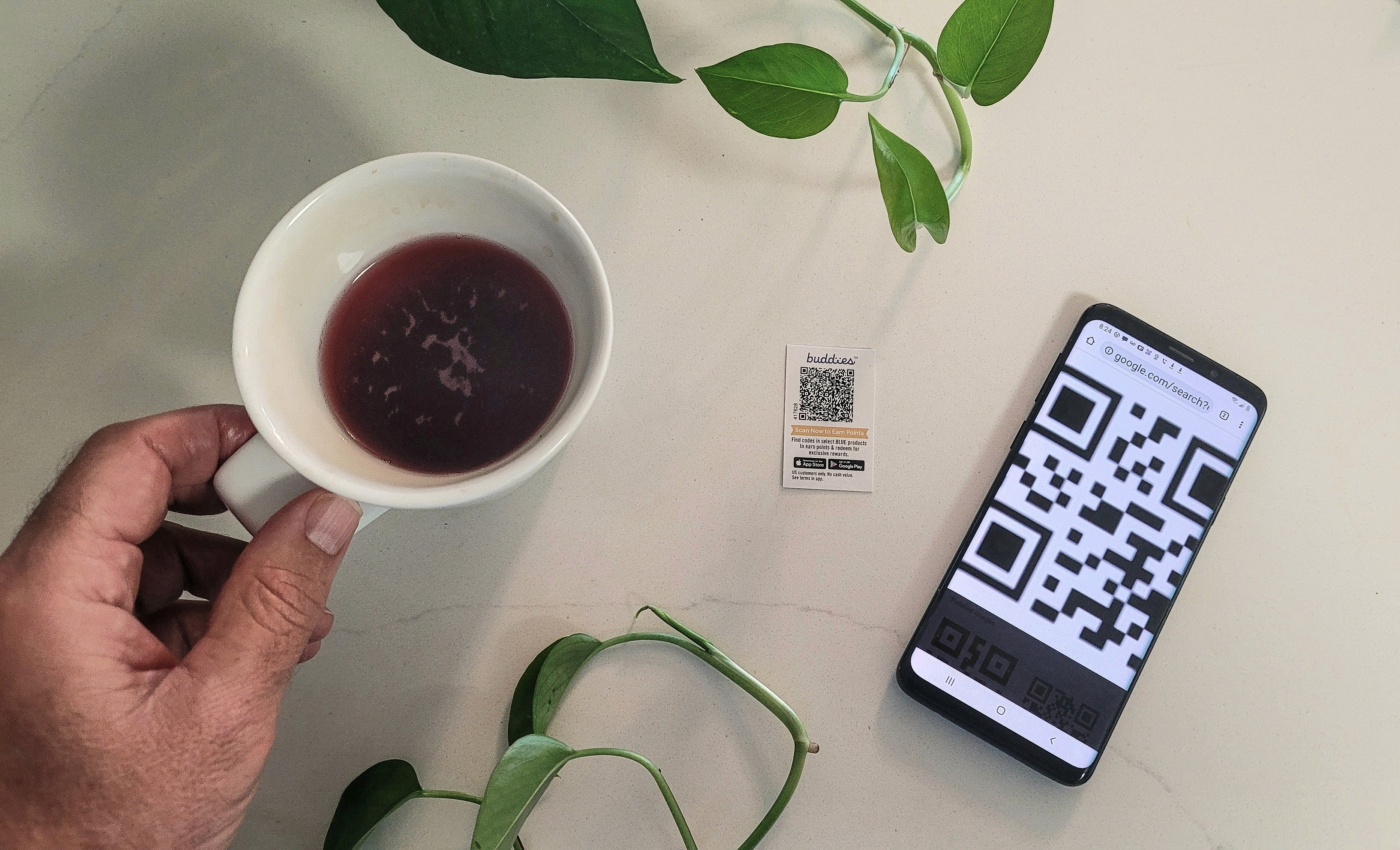 Eine Nahaufnahme zeigt eine Hand, die eine Kaffeetasse hält, während auf einem weißen Tisch eine kleine gedruckte Quittung mit QR-Code liegt. Daneben wird auf einem Smartphone-Display ein weiterer großer QR-Code angezeigt. Dies symbolisiert den Prozess der digitalen Rechnungsprüfung oder das Quishing-Risiko.