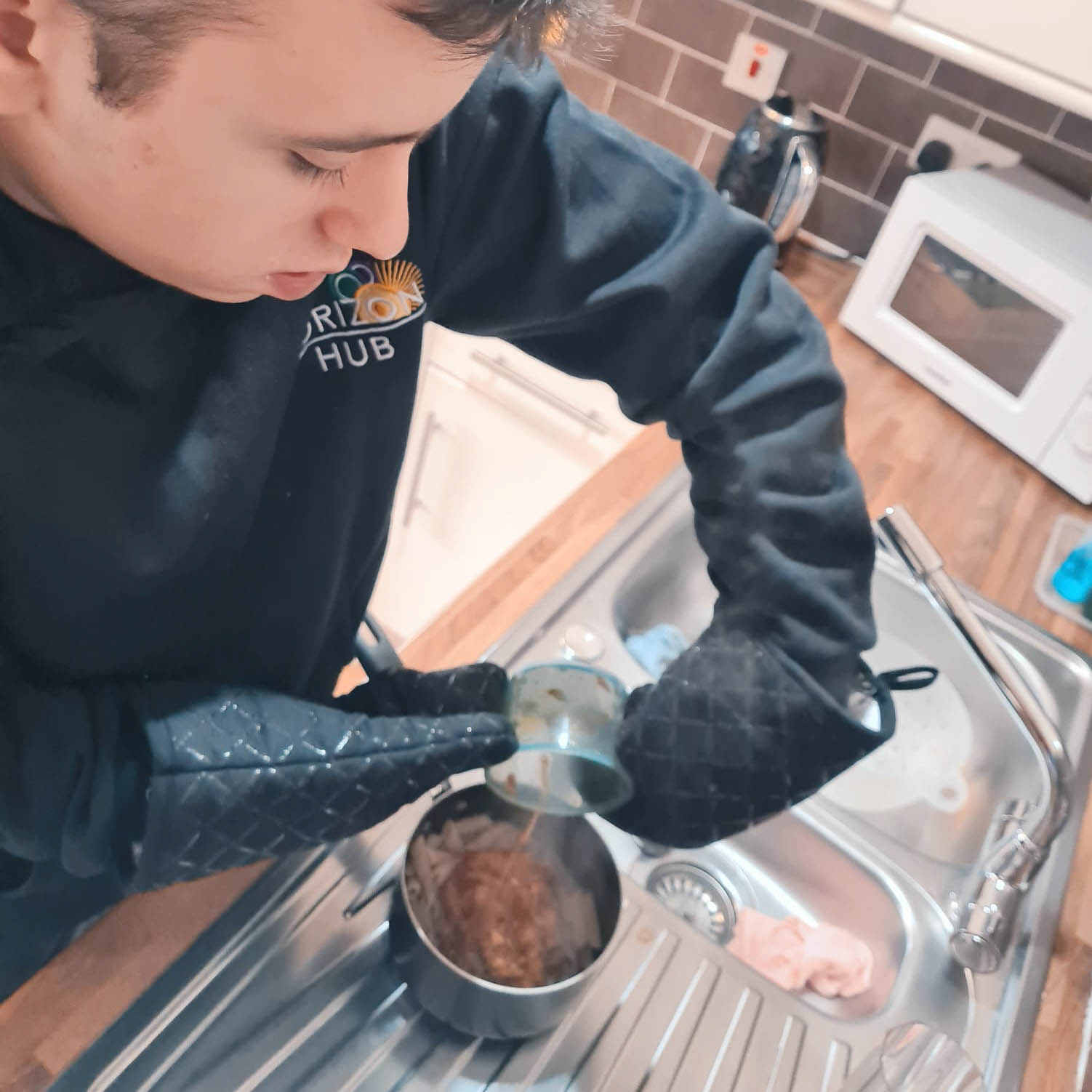 A teenager is preparing a meal using oven gloves and a saucepan