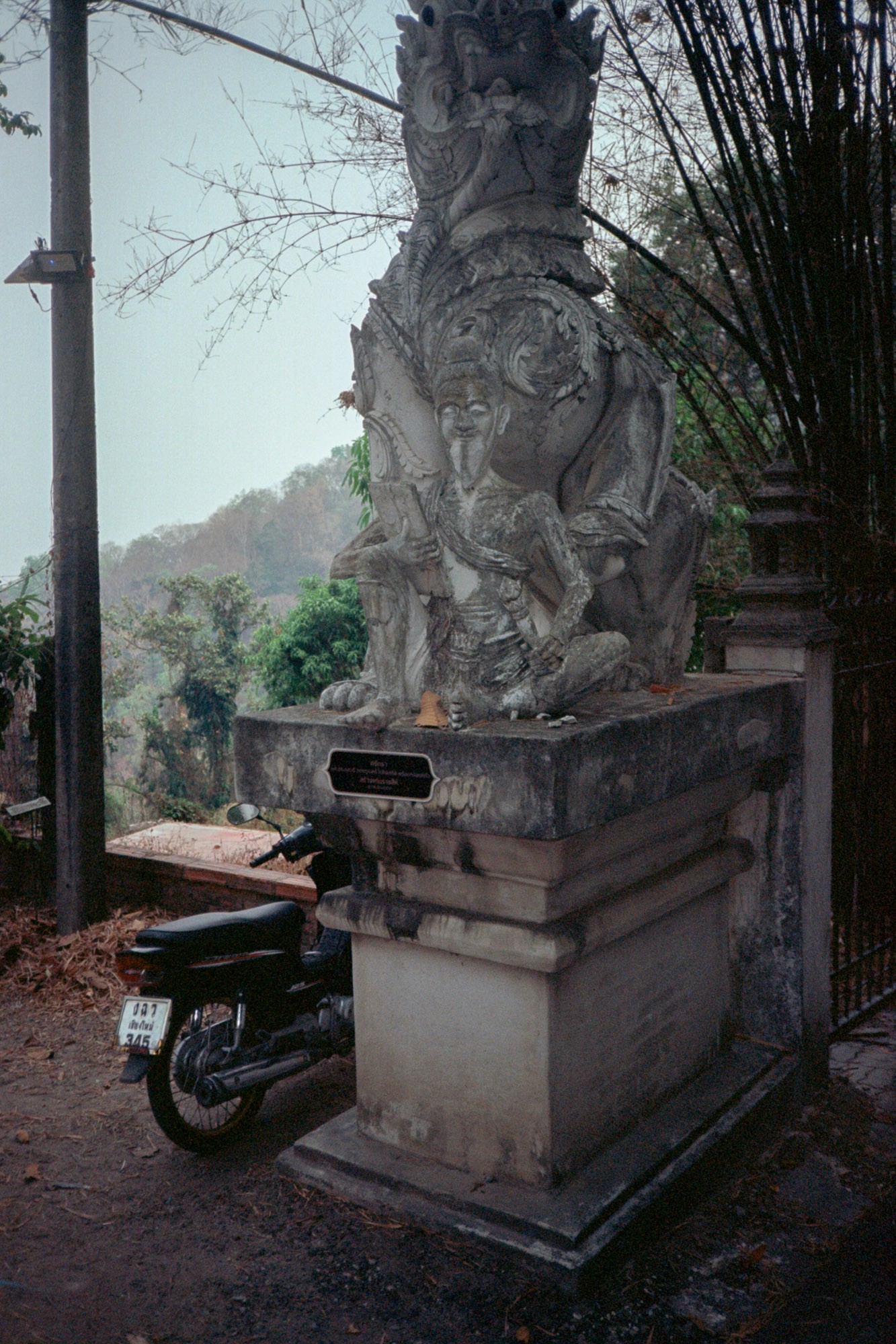 A stone sculpture of a mythical creature, possibly a guardian figure, is situated on a pedestal near a forested hillside, with a motorbike parked beside it, surrounded by bare trees and sparse vegetation.