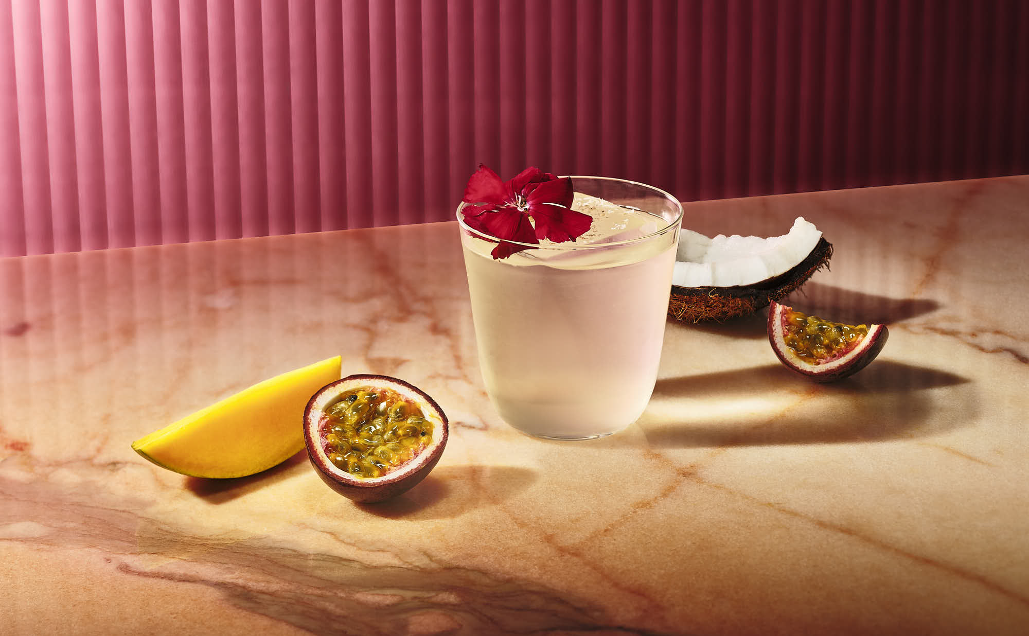 A white cocktail with red flower sits on a wooden surface, accompanied by mango slice and a soft, pink backdrop.