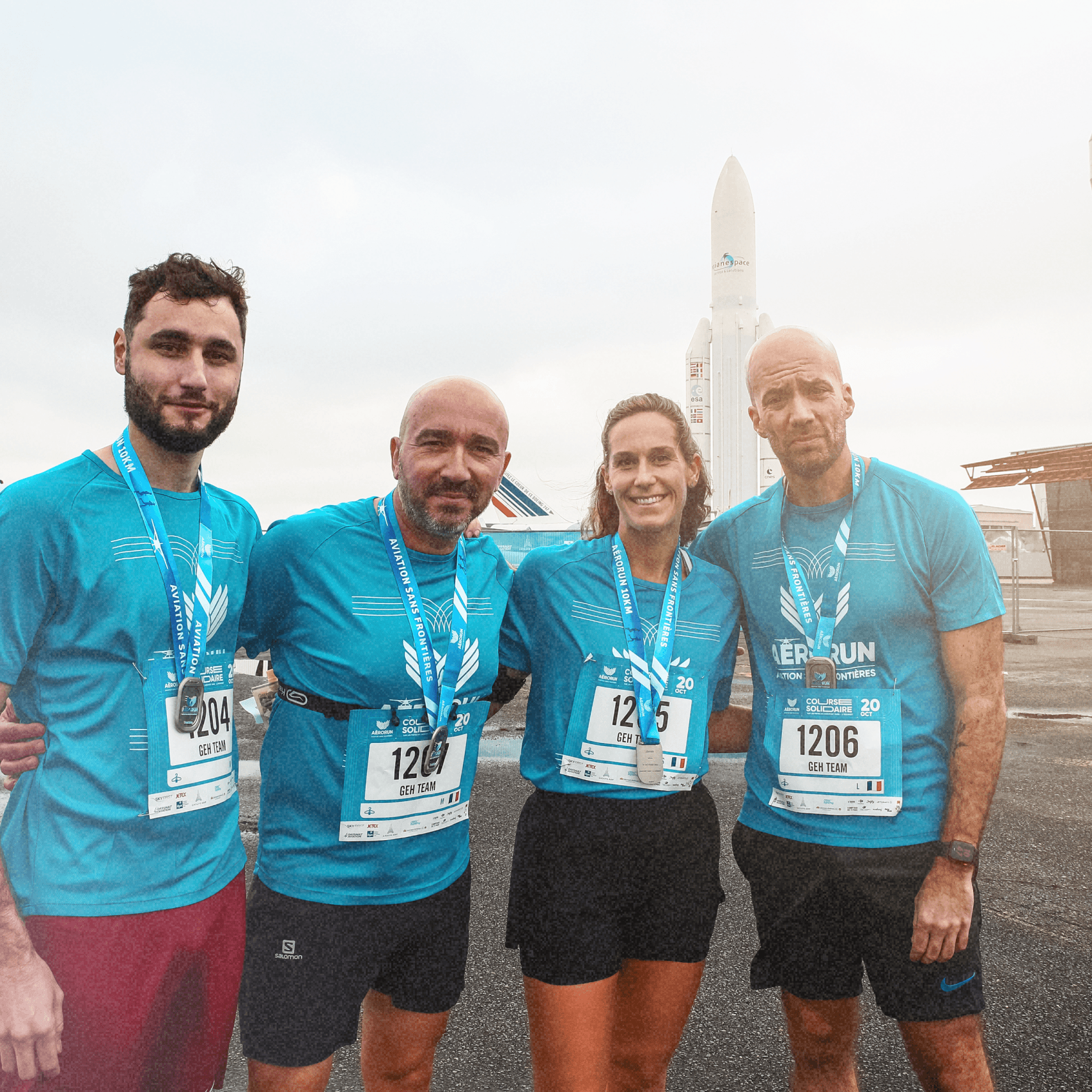 Groupe de quatre coureurs souriants avec des médailles autour du cou après avoir terminé une course, avec des fusées en arrière-plan.