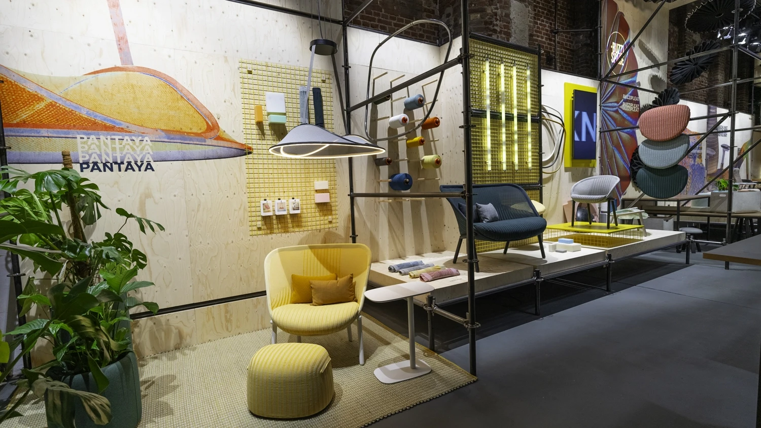 Booth area with yellow armchair, pouf, sample boards and colourful textile objects on scaffolding.