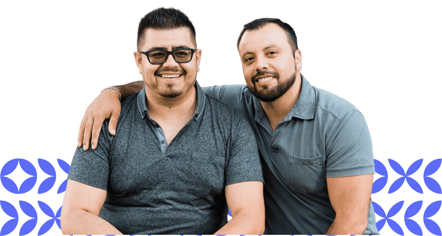 Two men are sitting close together, both smiling and wearing casual short-sleeved shirts, with one having his arm around the other's shoulder, against a transparent background with a subtle blue pattern.