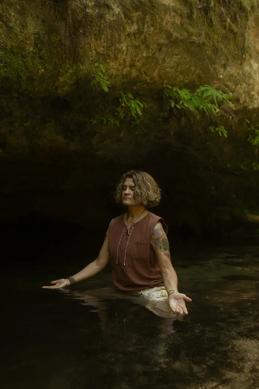 Sonia kneeling in a jungle stream with arms open in a posture of surrender and embodied presence