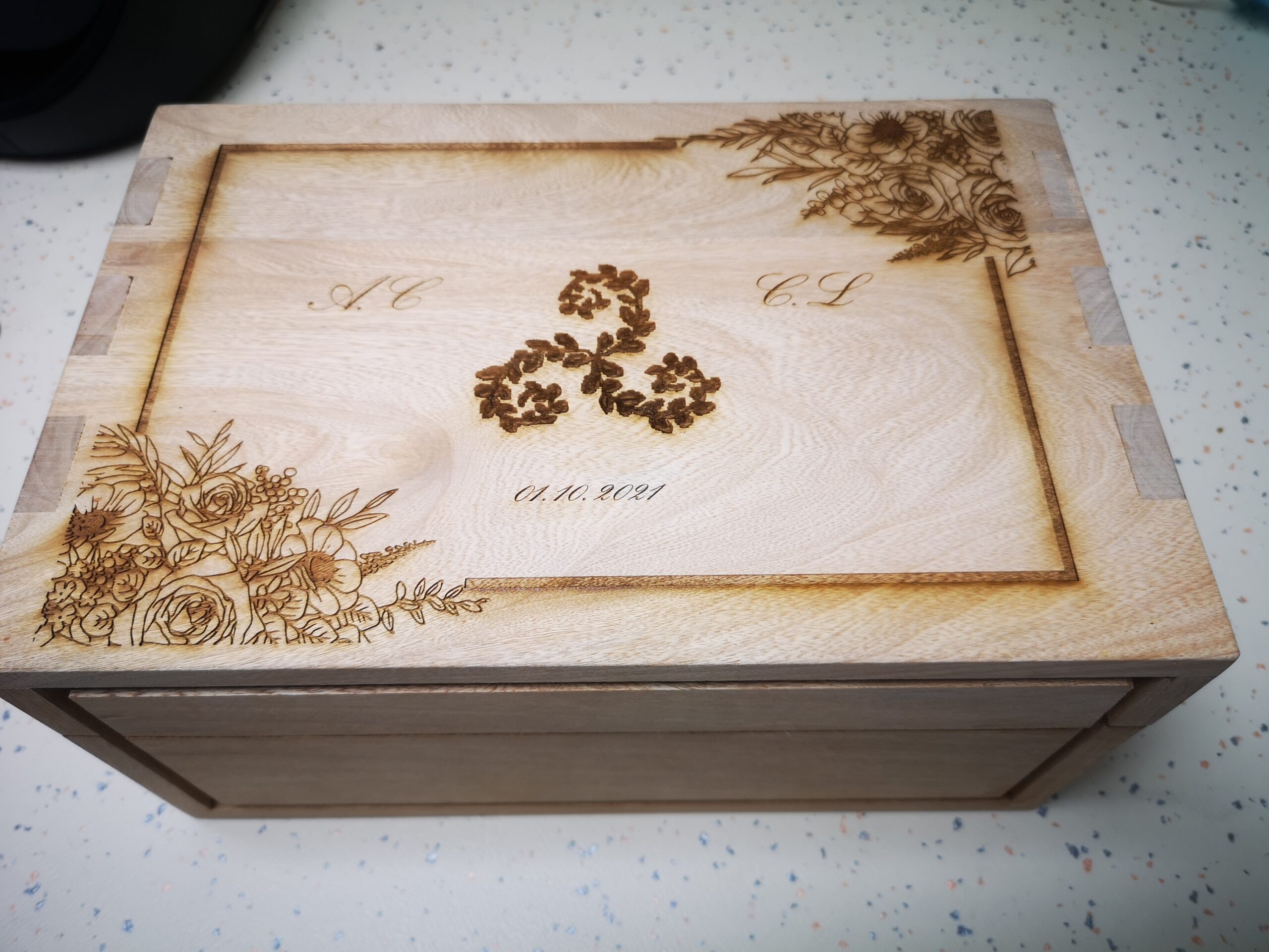 Boîte en bois avec des gravures florales sur les coins supérieurs et les initiales « A.C » et « C.L. » en écriture cursive. Motif floral central au-dessus de la date « 01.10.2021 ». Rustique et élégante.