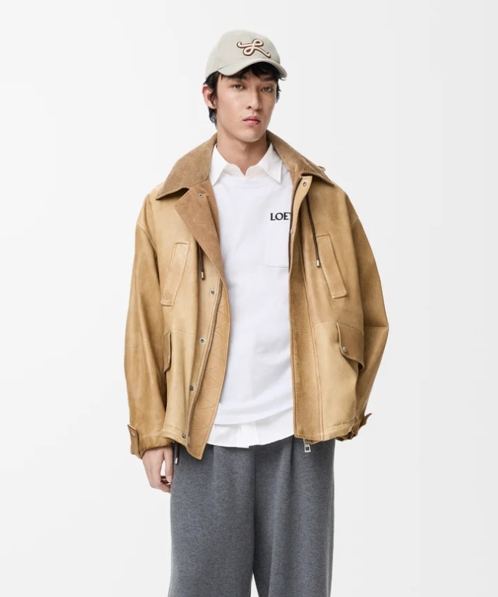 Smiling man in a white hoodie and black pants standing confidently on a bright green football field with empty stadium seats behind.