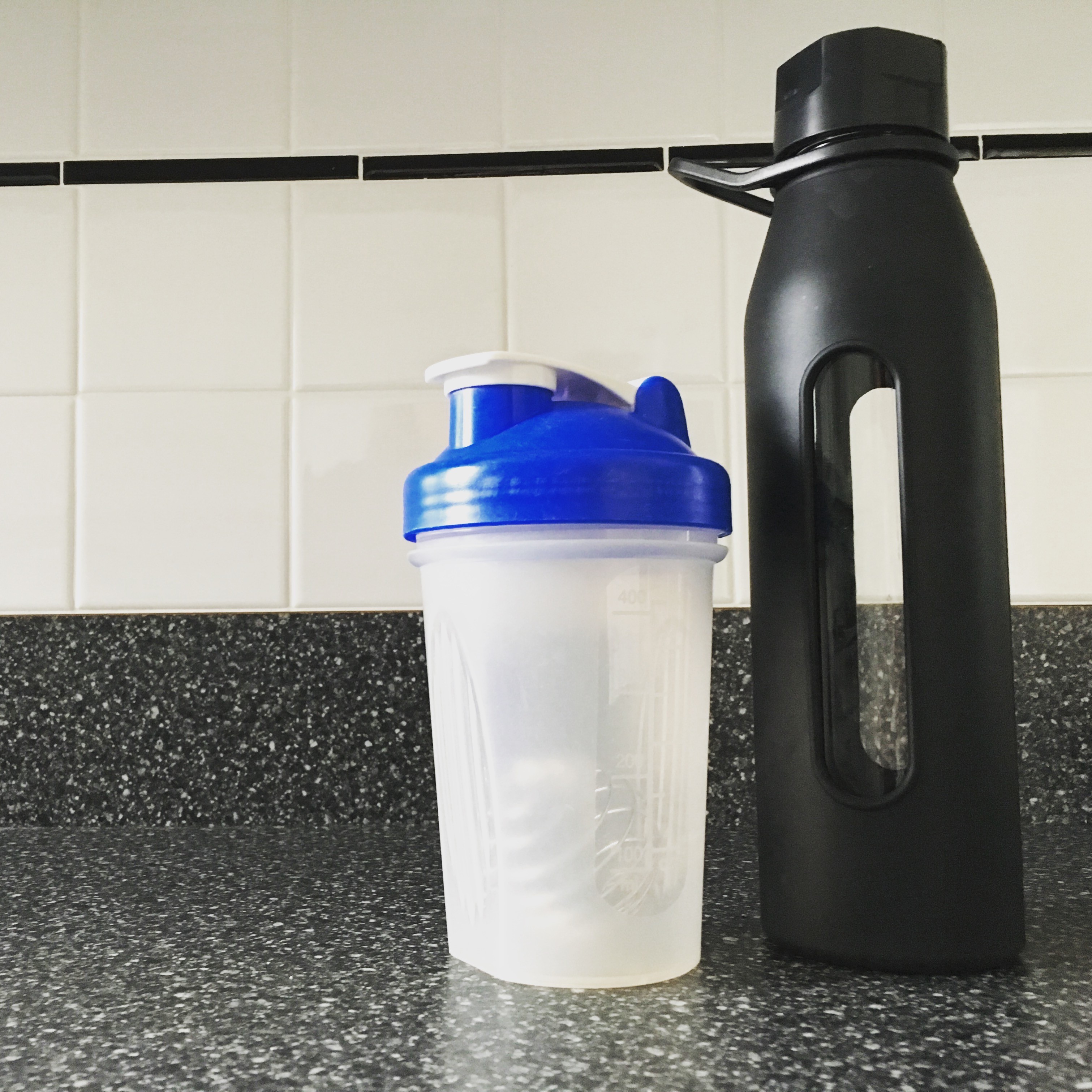 A white and blue shaker bottle and a black water bottle sit on a speckled granite countertop against a backdrop of white tiled walls.