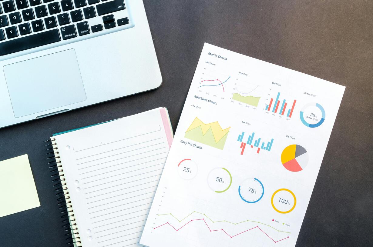 Laptop with financial charts and graphs, notebook, and a yellow sticky note on a dark desk.