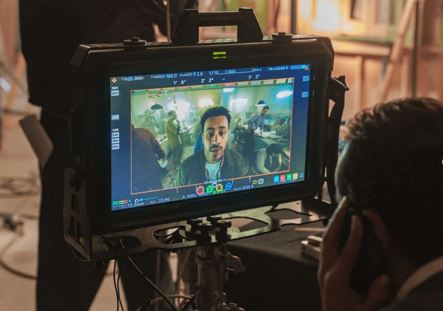 A person monitoring a film scene on a screen, with various lights and crew members in the background.