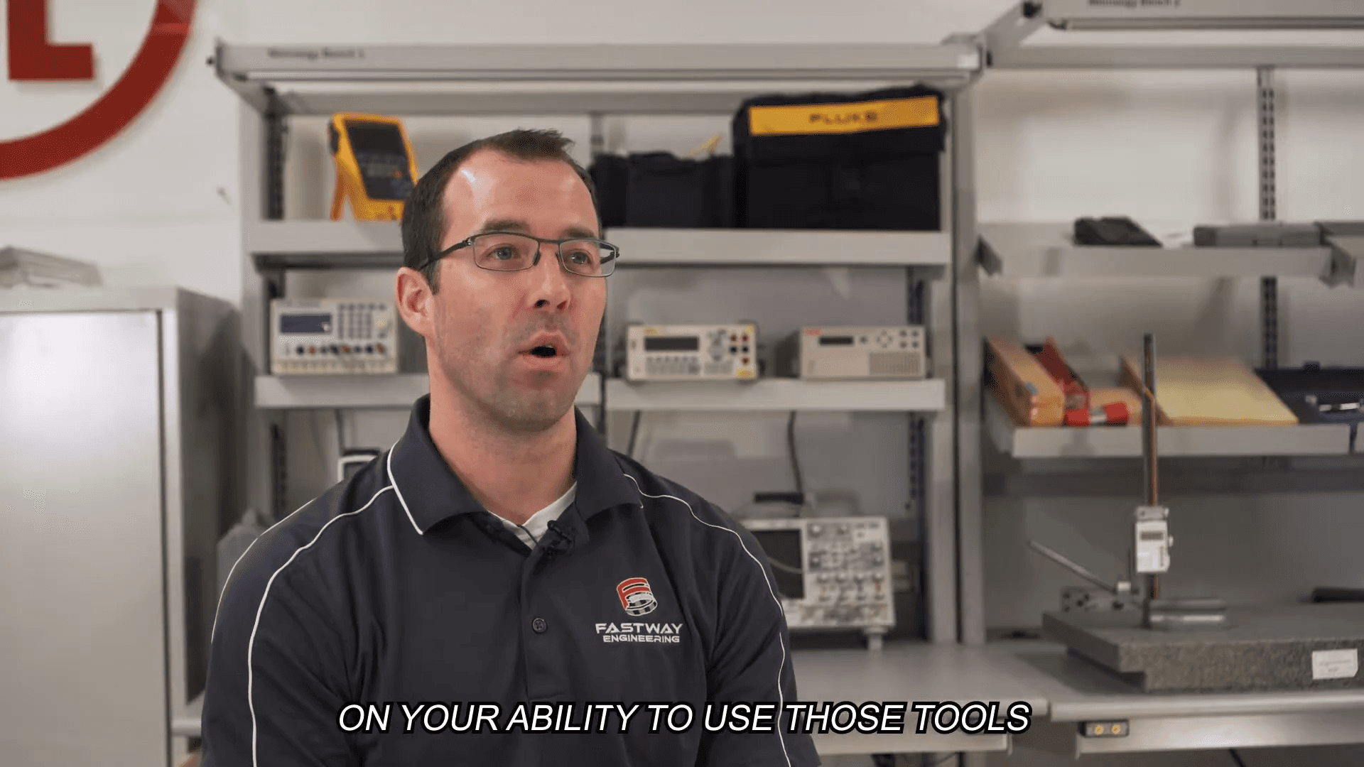 A professional in a workshop or lab setting speaking to the camera, with tools and equipment visible in the background.
