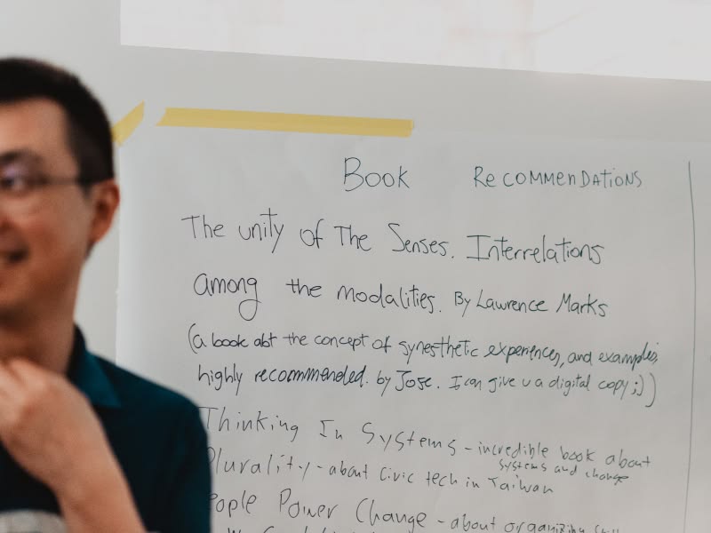 A person smiling on the left, standing beside a whiteboard with handwritten book recommendations. The tone is informal and engaging.