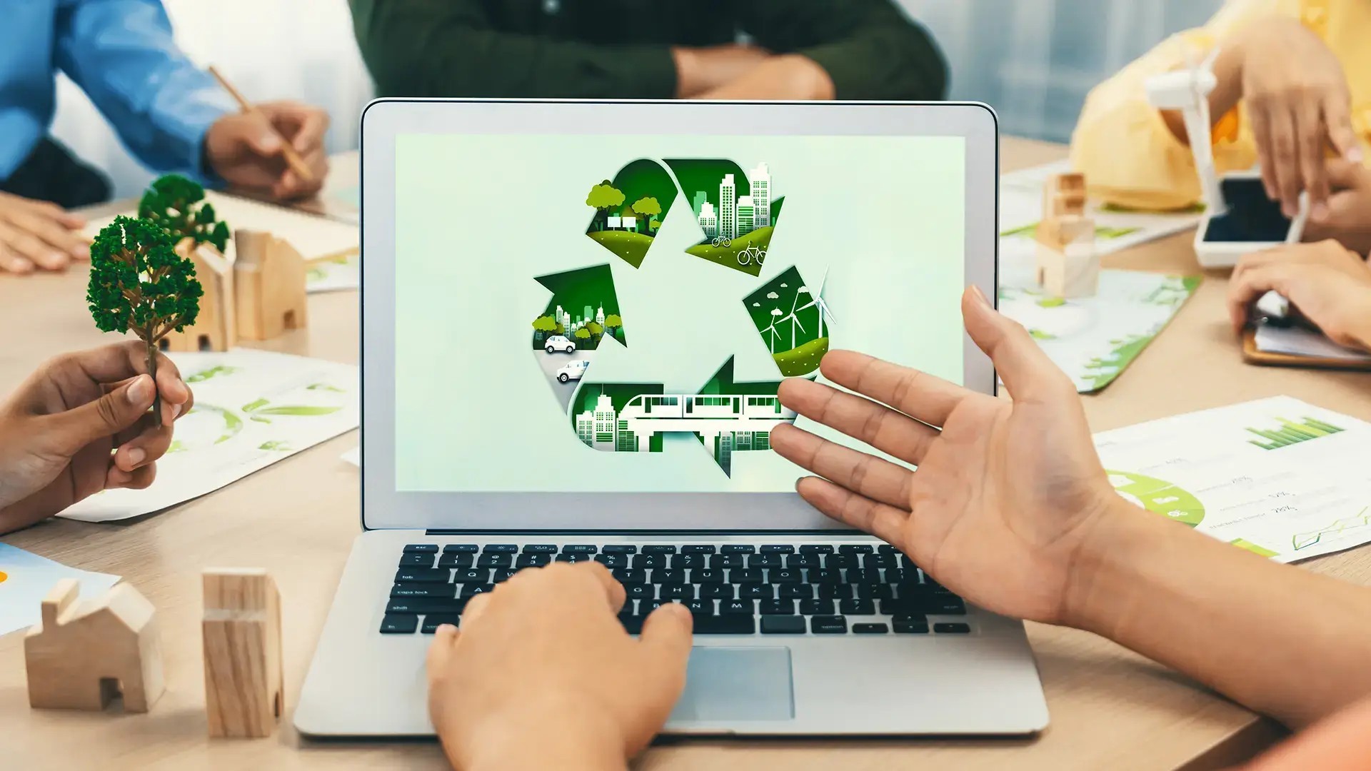 People sat at a table grouped around a laptop showing a green recycling logo with images of infrastructure within it. One person’s hand is gesturing towards the logo, symbolising a corporate discussion on green initiatives and strategies for the global future.