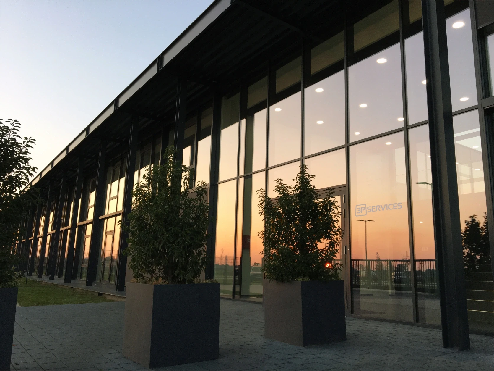 Modern headquarter building with large windows reflecting sunset colors and potted plants outside the entrance.