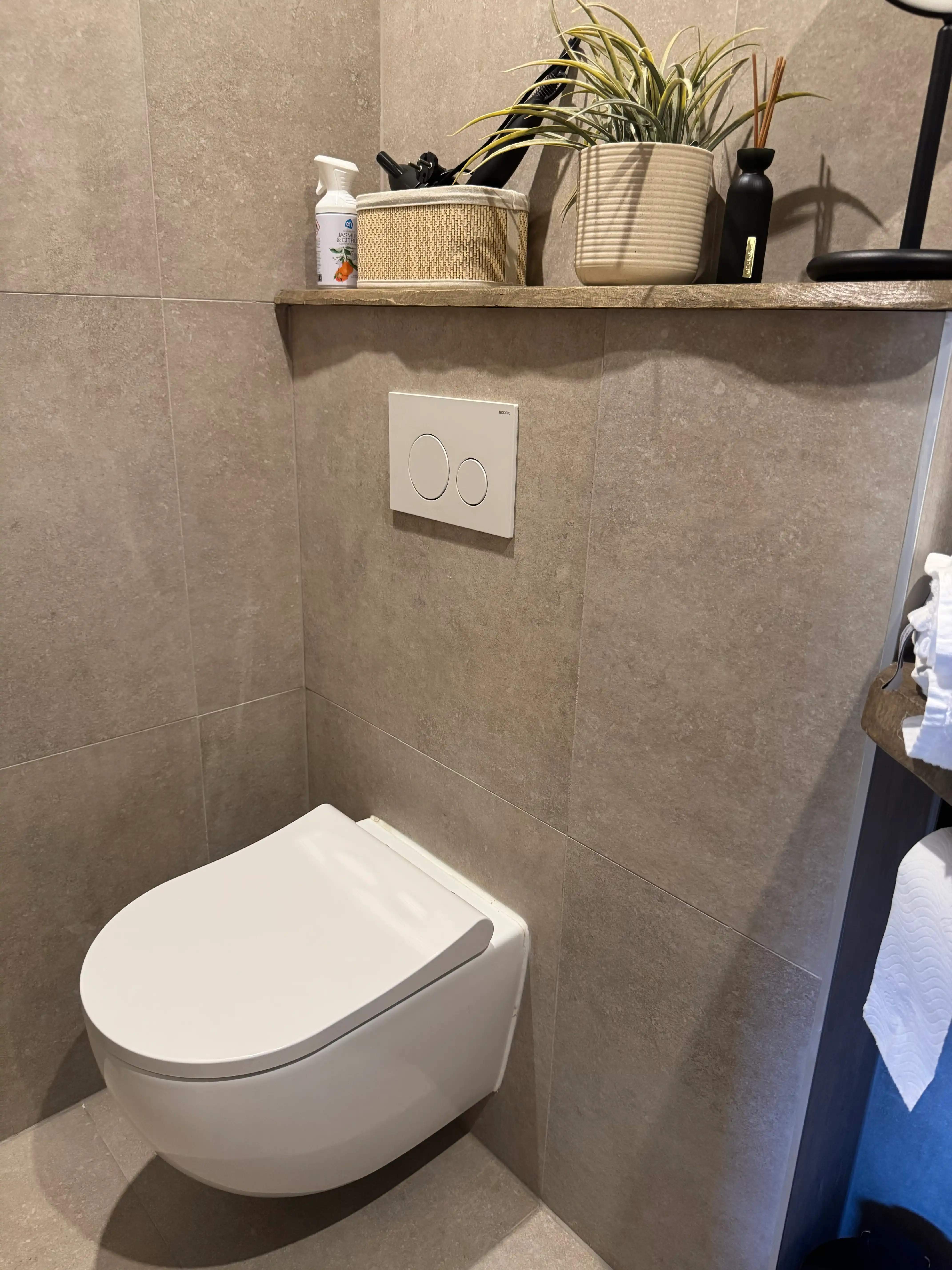 Modern zwevend toilet met beige tegels en houten wandplank.