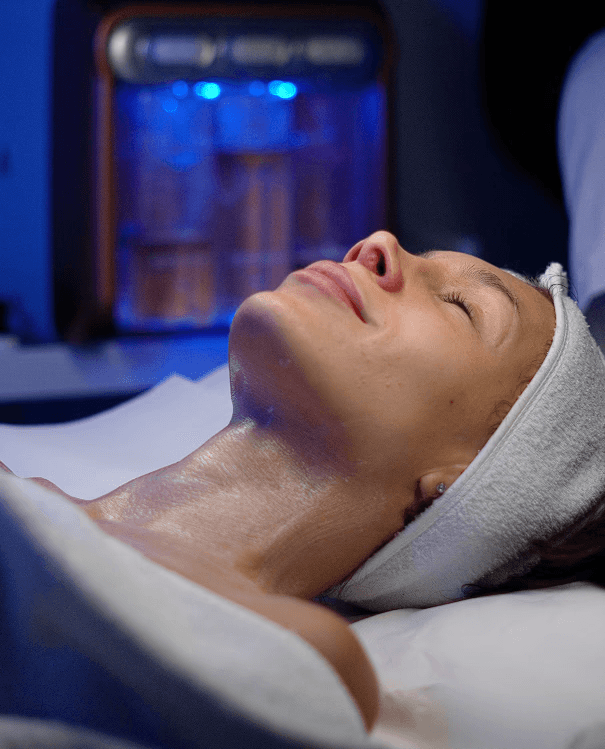 Woman in a towel and head wrap applying skincare cream in a cozy bedroom