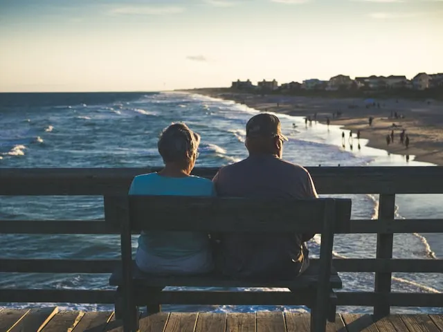 Pareja mayor sentada de espaldas en un banco de un paseo marítimo, contemplando la playa al atardecer, representando la jubilación que planifica un seguro de vida ahorro.