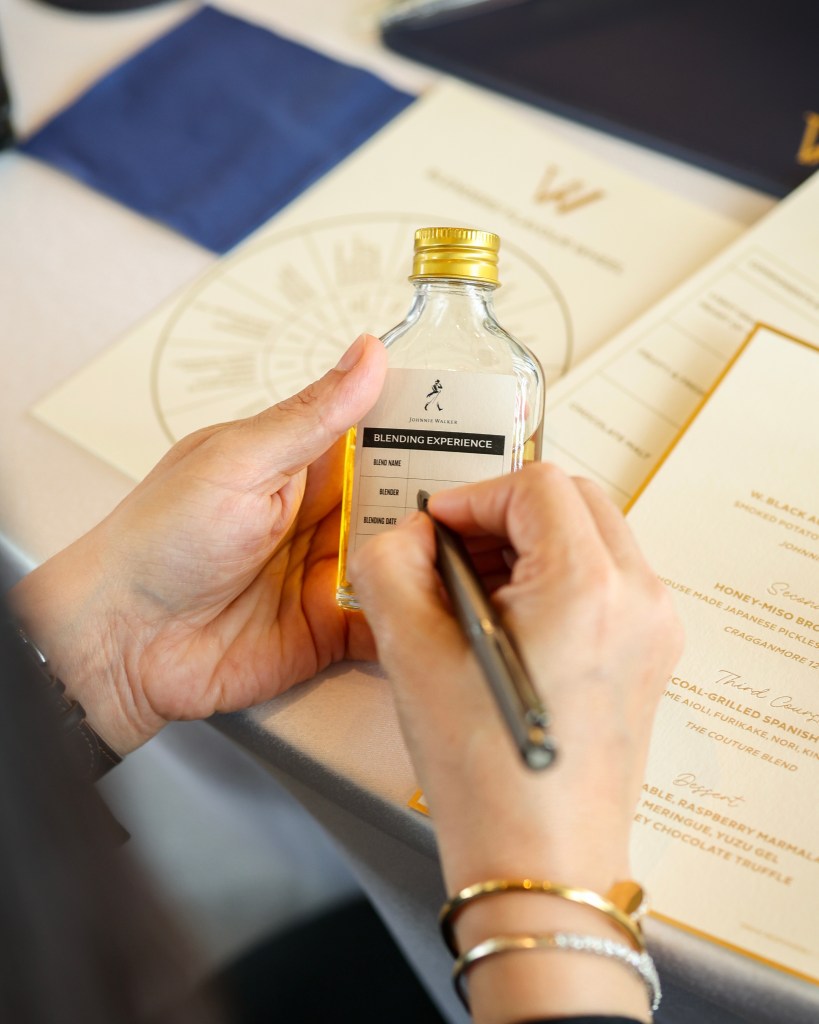 A person holding a small glass bottle labeled 'Blending Experience' while writing on a label. The background shows a neatly arranged table setting with a menu.