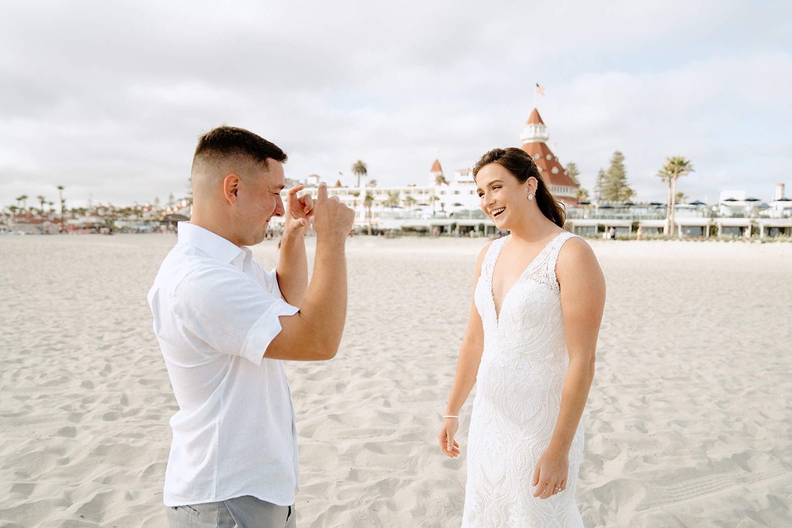 A man photographing a woman in a flowing dress on a sandy beach at Hotel Del Cornado, with ocean and palm trees in the background.