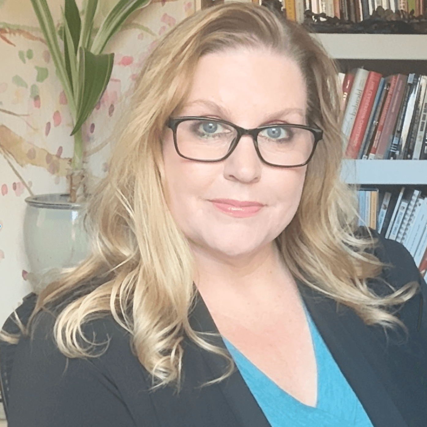 A woman with glasses and wavy blonde hair smiles subtly. She's in an office with bookshelves and a potted plant, creating a warm, professional atmosphere.
