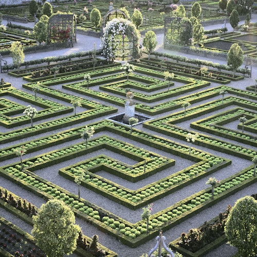 An ornate garden maze with trimmed hedges, topiary trees, flower beds, and a central statue under a sunny sky.