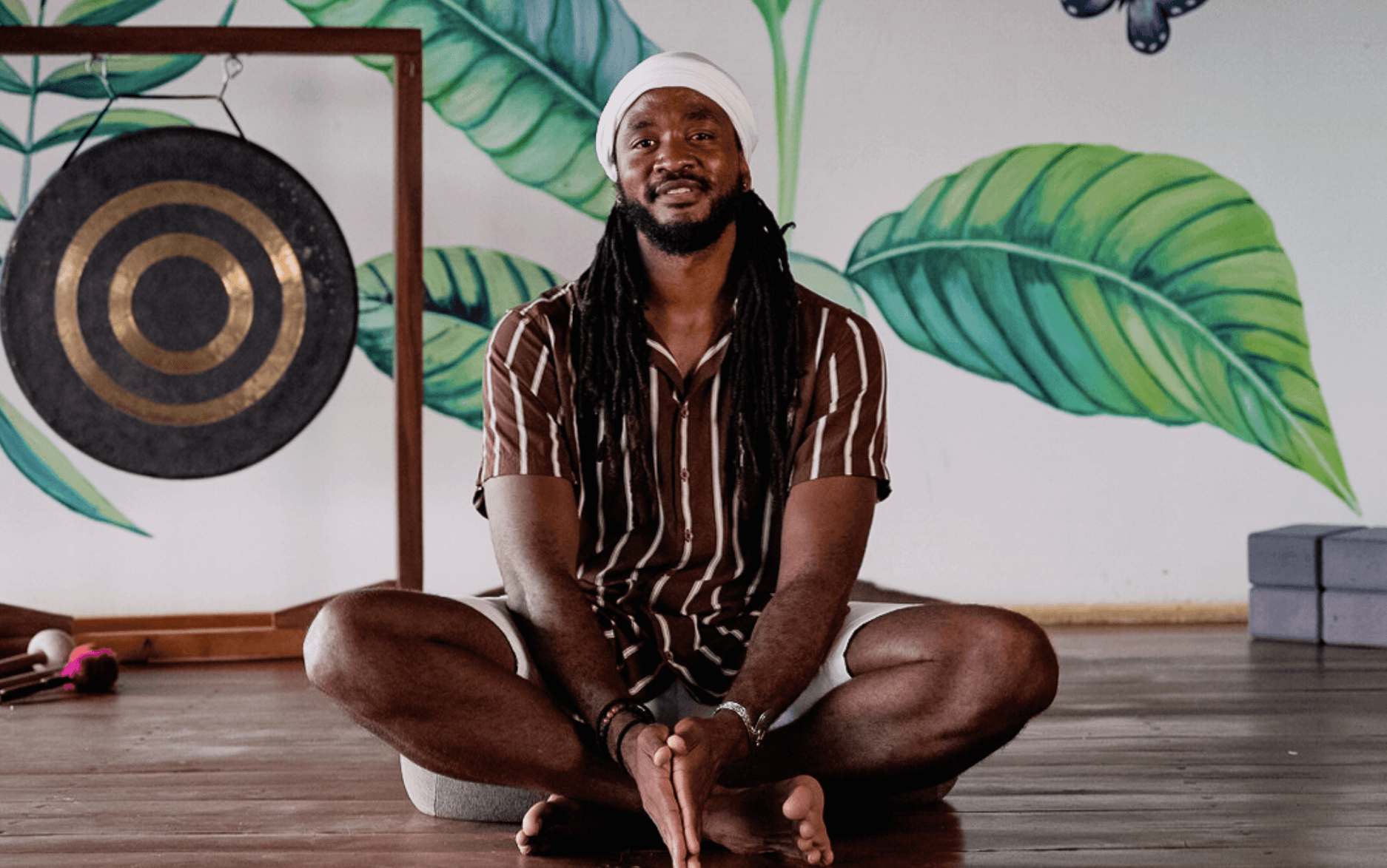 Confident man standing in a greenhouse surrounded by lush green plants, wearing a beige shirt and neutral-toned outfit.