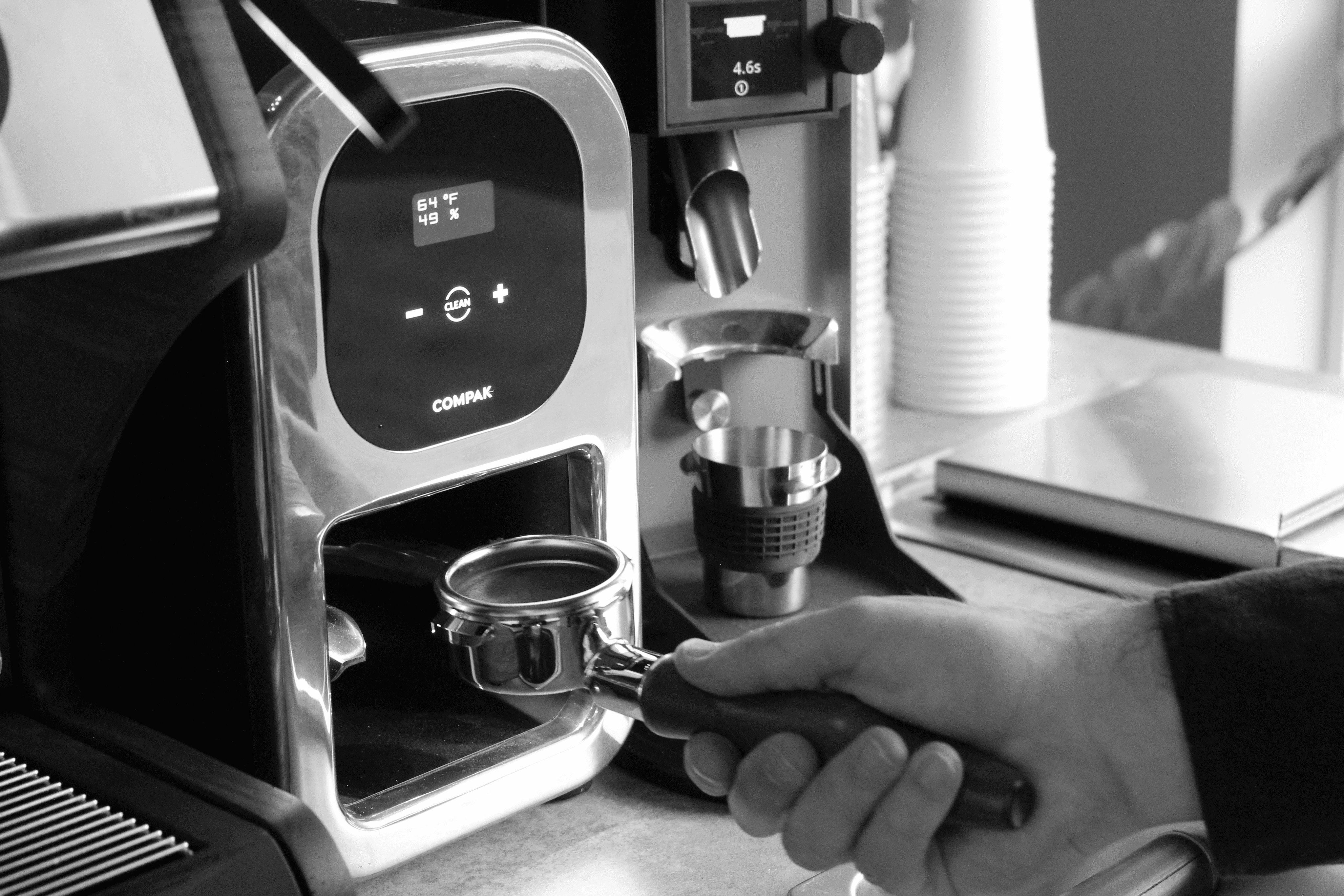 Black and white close-up of a person holding a portafilter under a digital coffee grinder labeled “COMPAK.” The grinder's display shows a temperature of 64°F and humidity of 49%, with plus, minus, and clean buttons. Coffee grounds are being dispensed into the portafilter. In the background, a stainless steel dosing cup and a stack of paper cups are visible on the countertop. The image captures a precise moment in the espresso preparation process.
