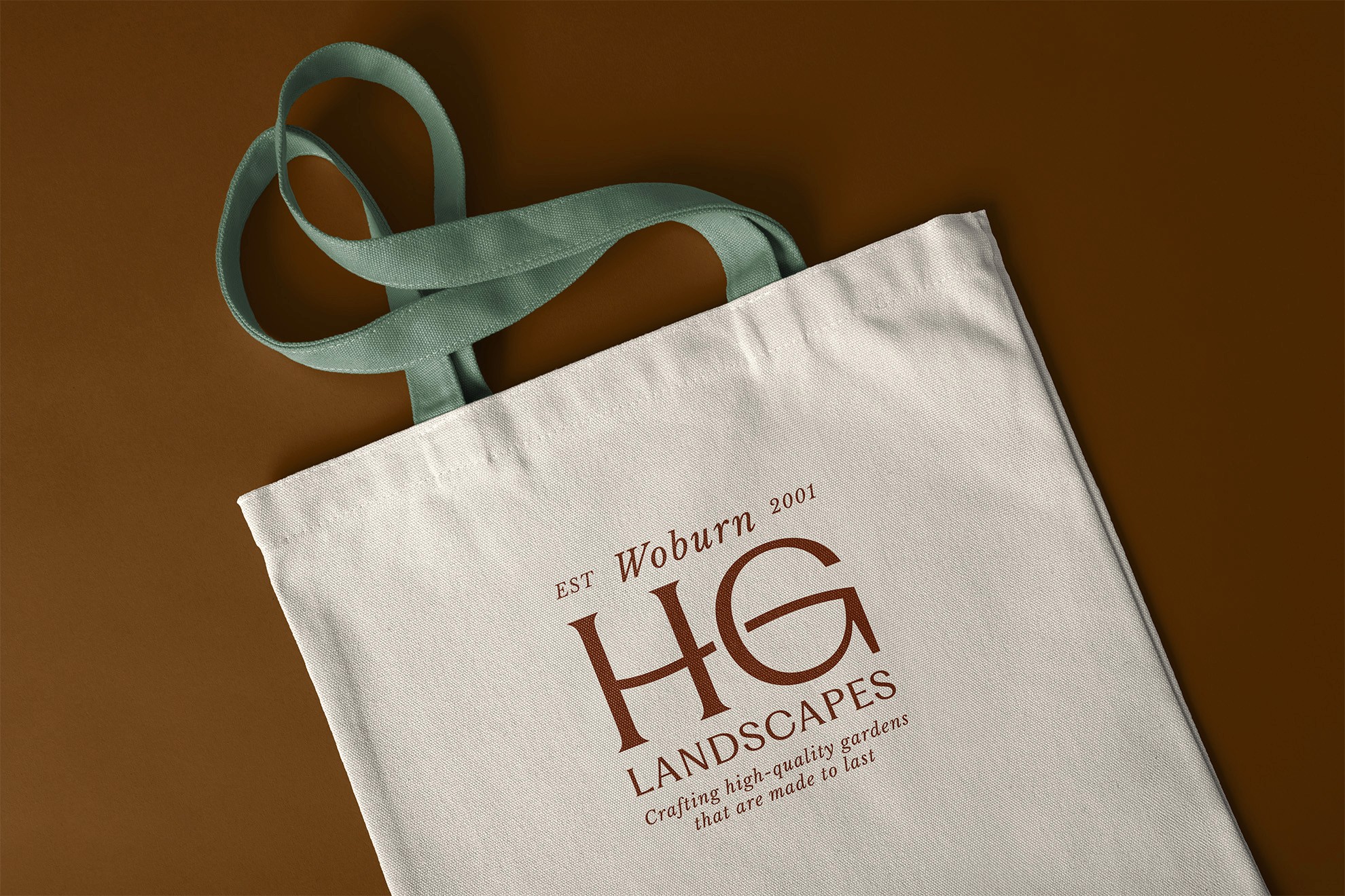 A white shopping bag with a teal handle and the letters "HG" printed on it, placed on a brown surface.