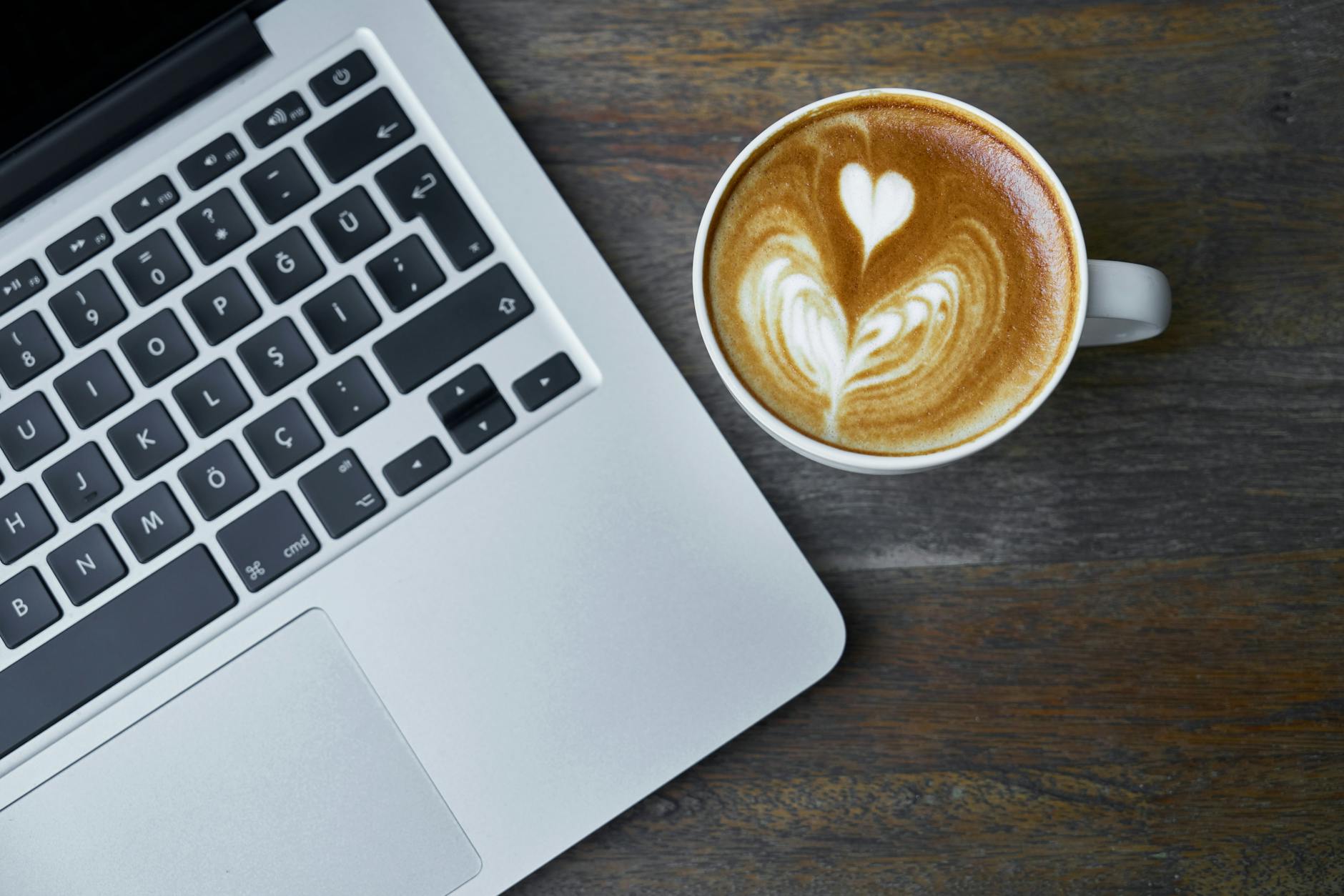A close-up of a wooden desk featuring a laptop, a steaming cup of coffee, and a stack of graded notebooks.