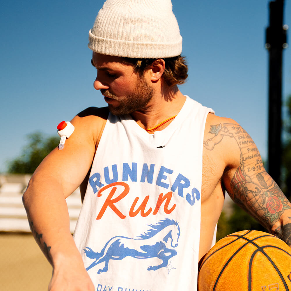 Man outdoors wearing a sleeveless shirt and beanie, looking at a Rythm Health device attached to his upper arm while holding a basketball.