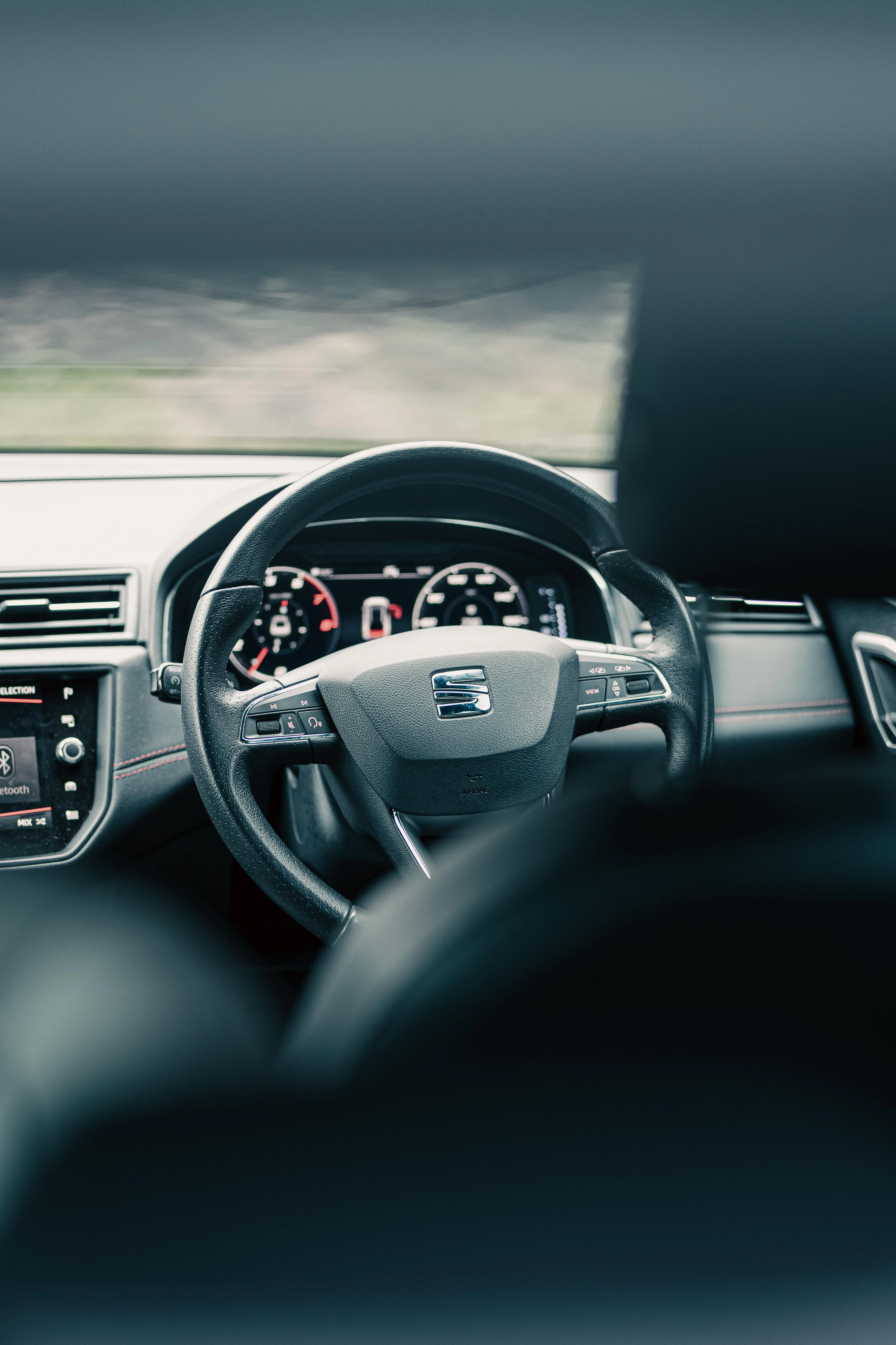 Vue centrée sur le volant SEAT et le tableau de bord, avec un léger flou en premier plan. L’image met en valeur une ambiance intérieure moderne, des commandes ergonomiques et une instrumentation claire, révélant le caractère fonctionnel et sportif de l’habitacle.