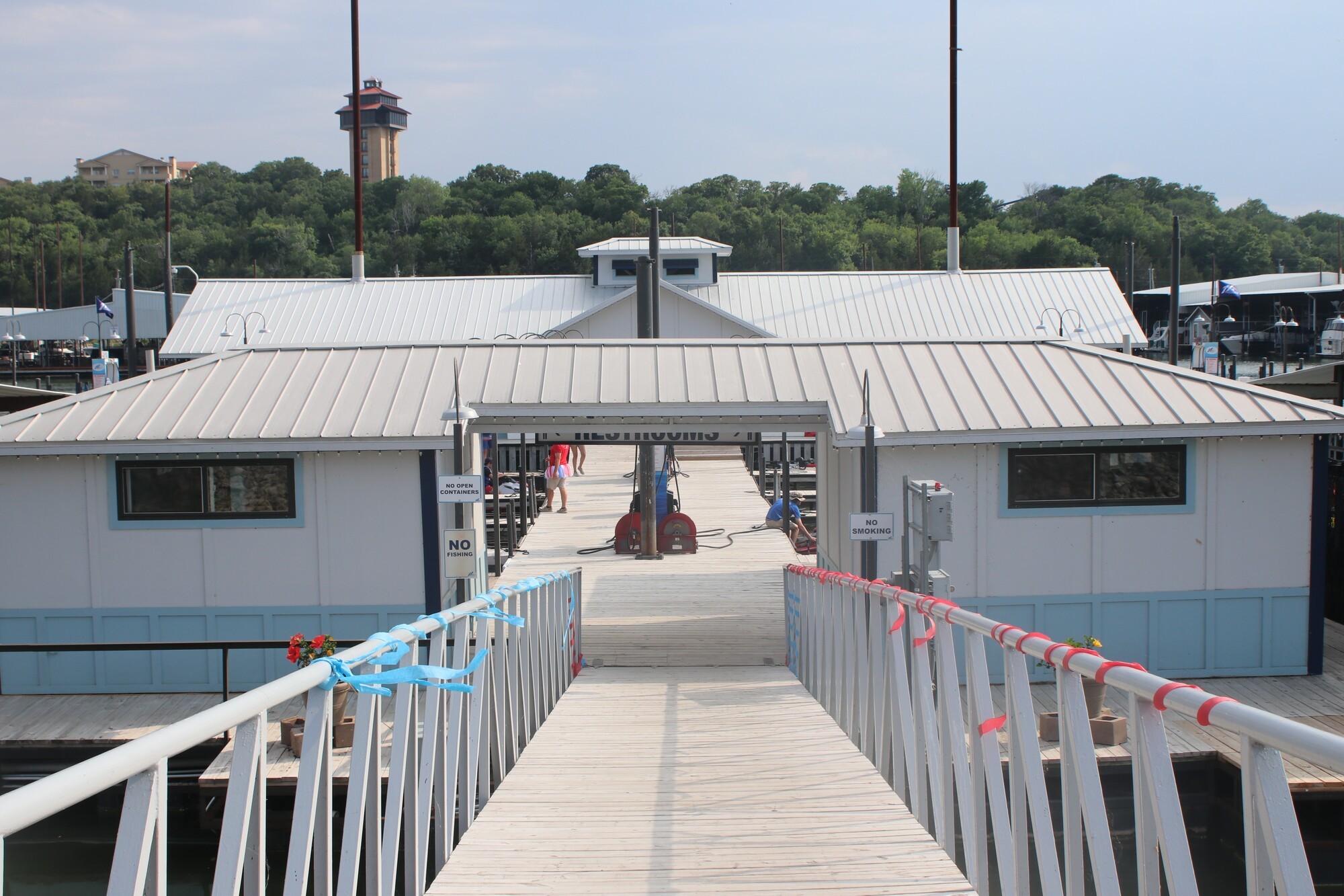 A wooden walkway adorned with colorful ribbons leads to a large, modern floating building at a marina, surrounded by lush green trees and a partly cloudy sky.
