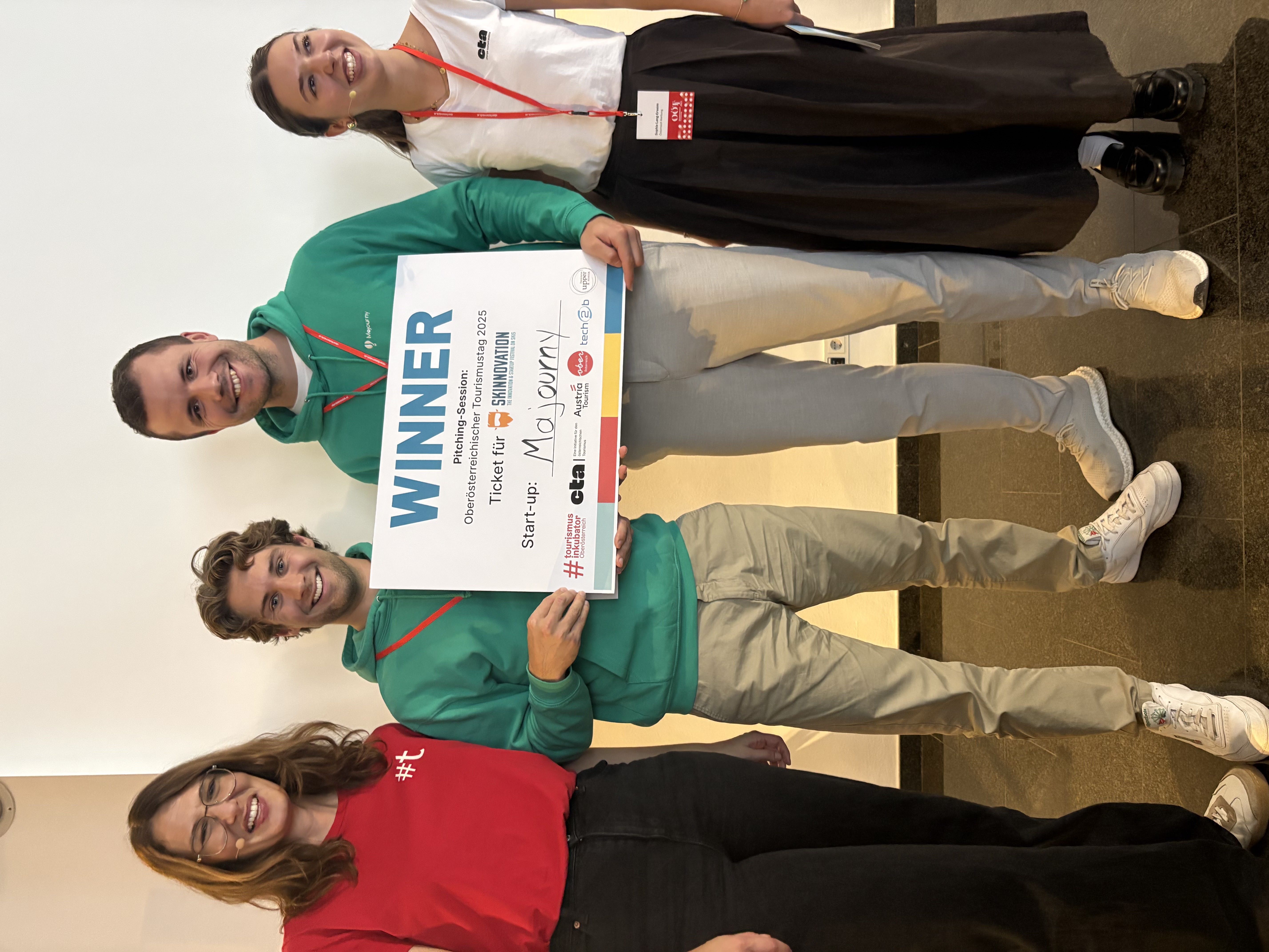 A group of four people stand smiling, with two men in green hoodies holding a "Winner" sign and flanked by two women, one in a red sweater and one in a black skirt, against a plain white background.