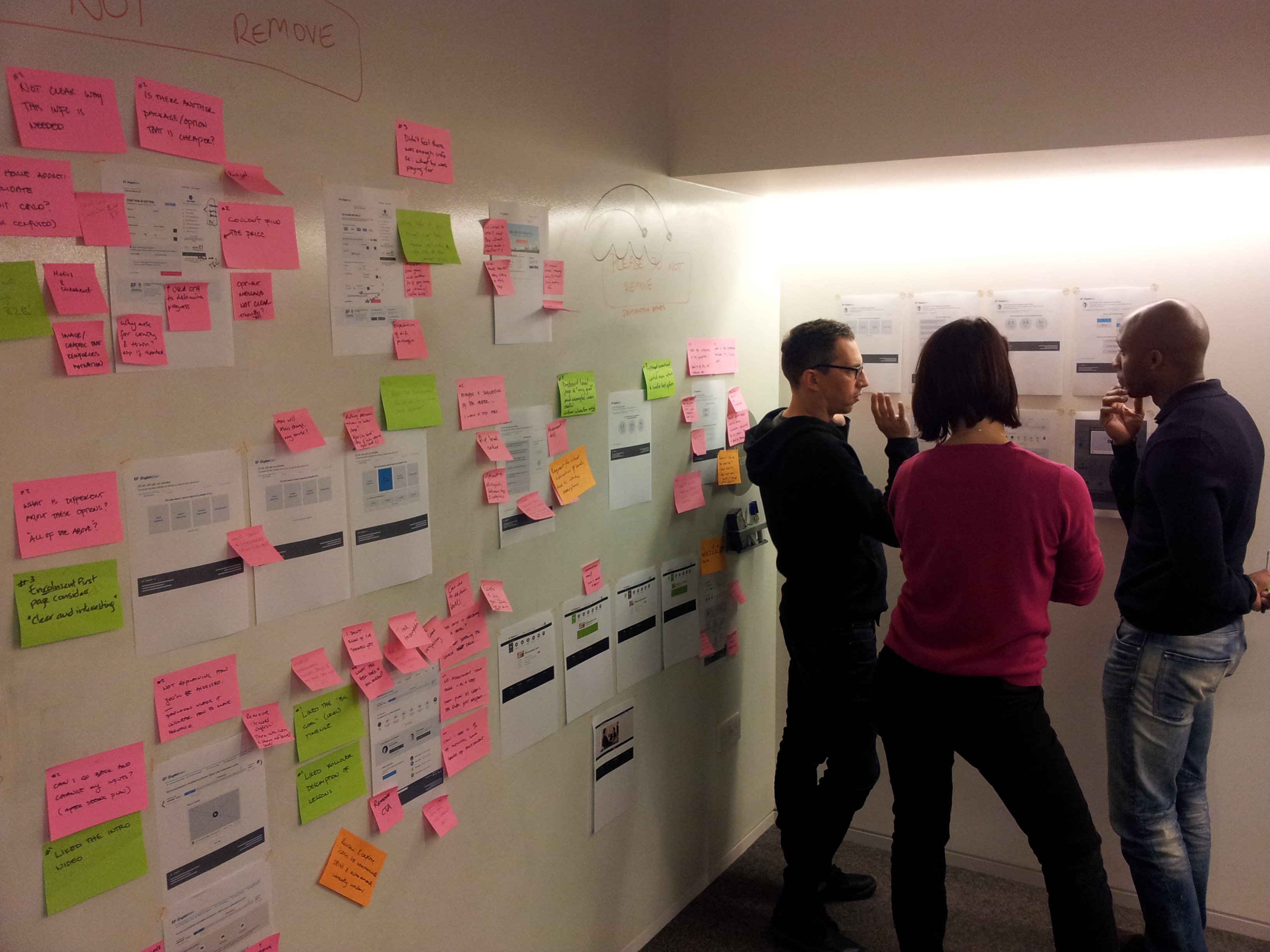 Three people standing at a wall discussing and looking at print-outs of wireframes. The wireframes are annotated with post-its.