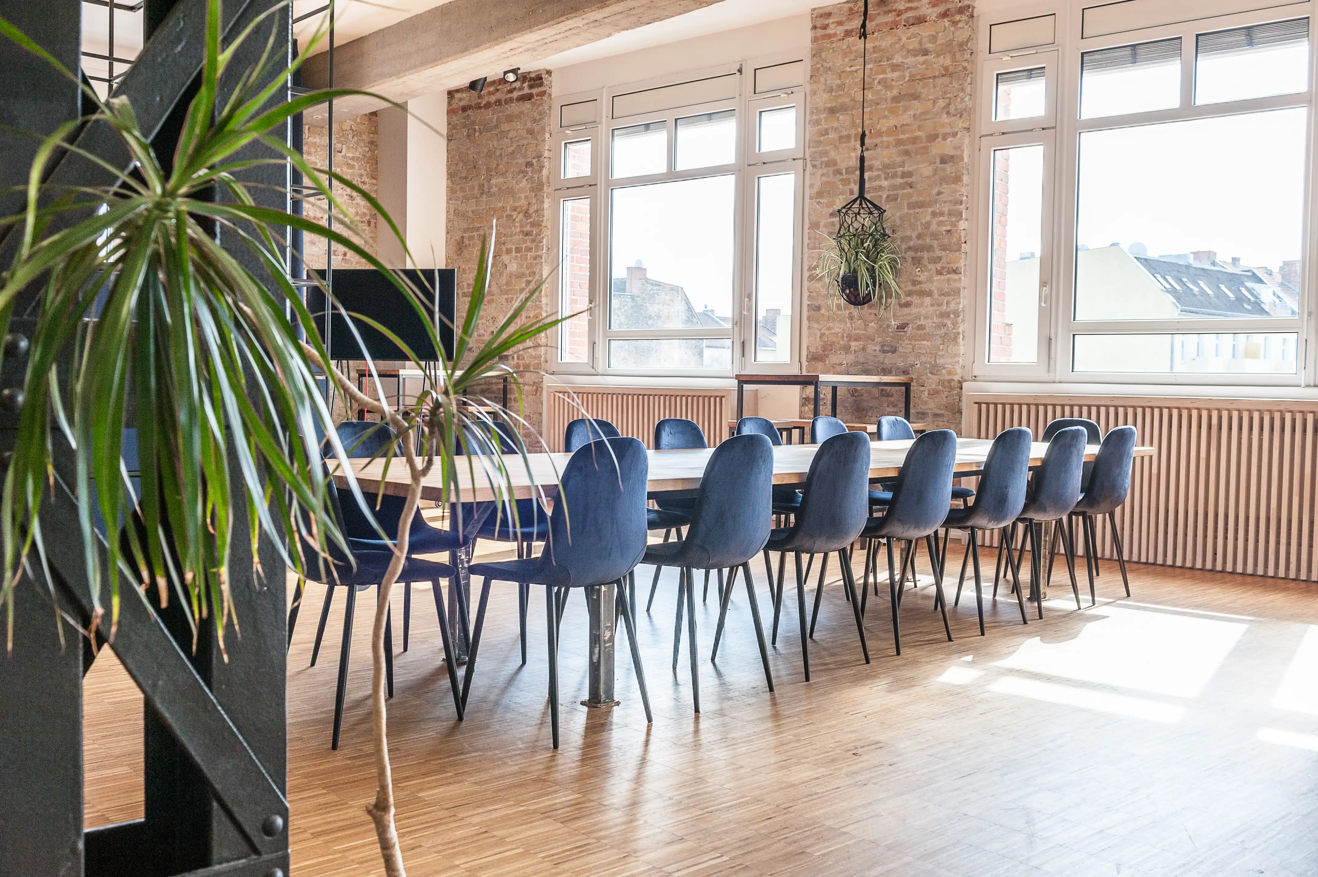 Long meeting table at BASILICOM with dark blue velvet chairs in front of exposed brick walls, white casement windows, a slatted wooden radiator cover and a hanging plant.