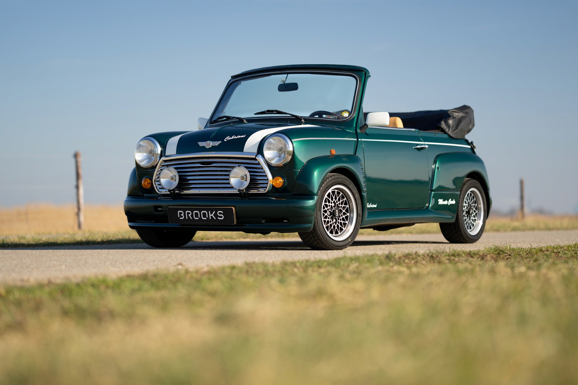 A vintage green mini classic automatic car drives along a road, surrounded by fields and a clear blue sky in the background.