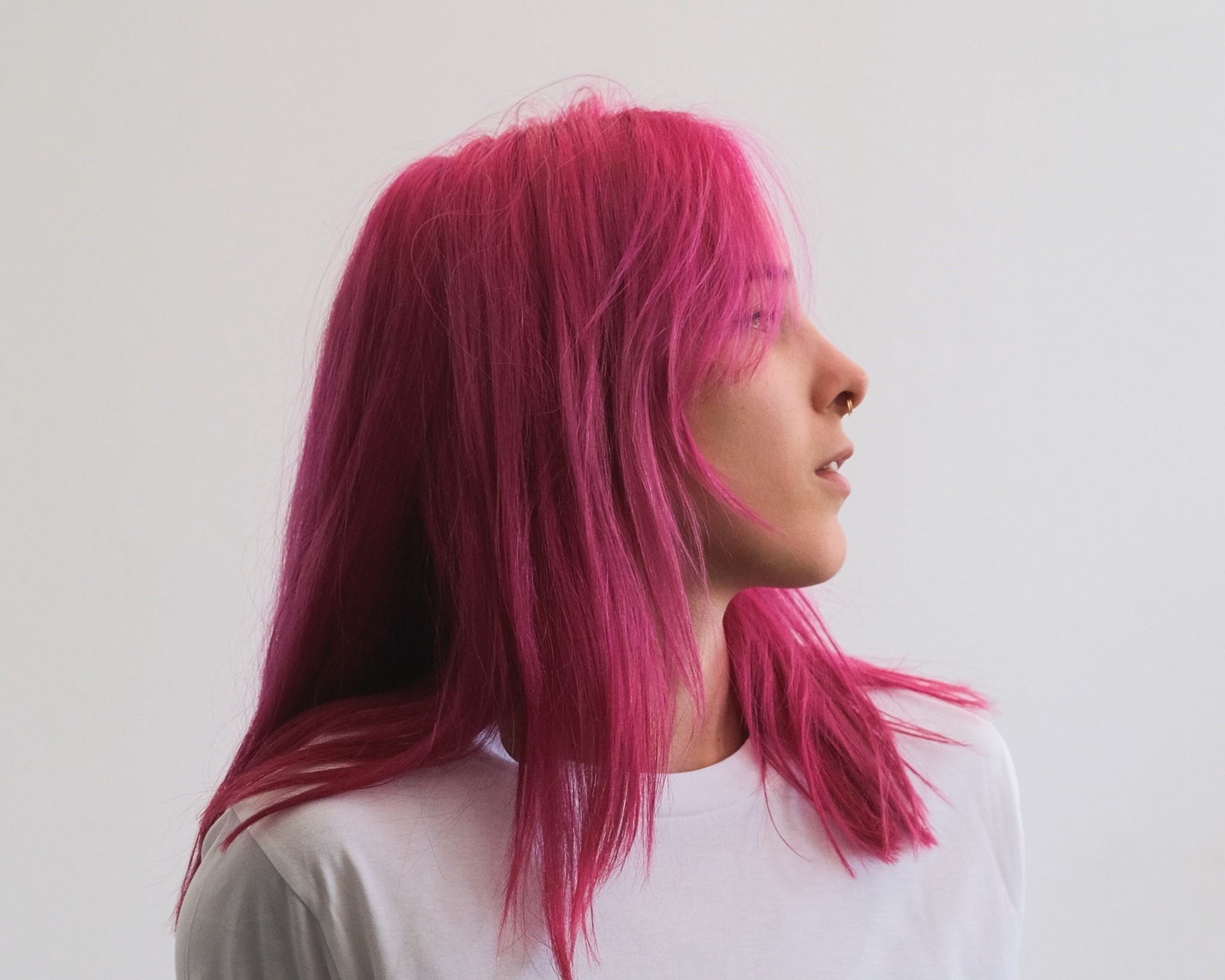 A white woman with pink hair stands against a white wall. She is in profile, facing left.