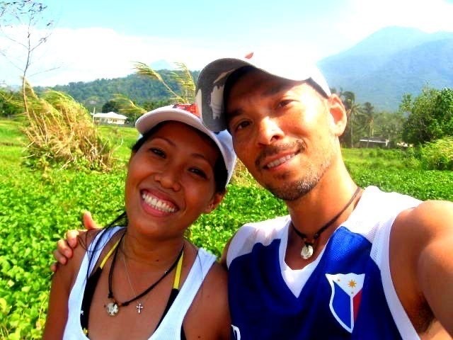 A smiling man and woman wearing hats posing for a selfie in a green tropical field.