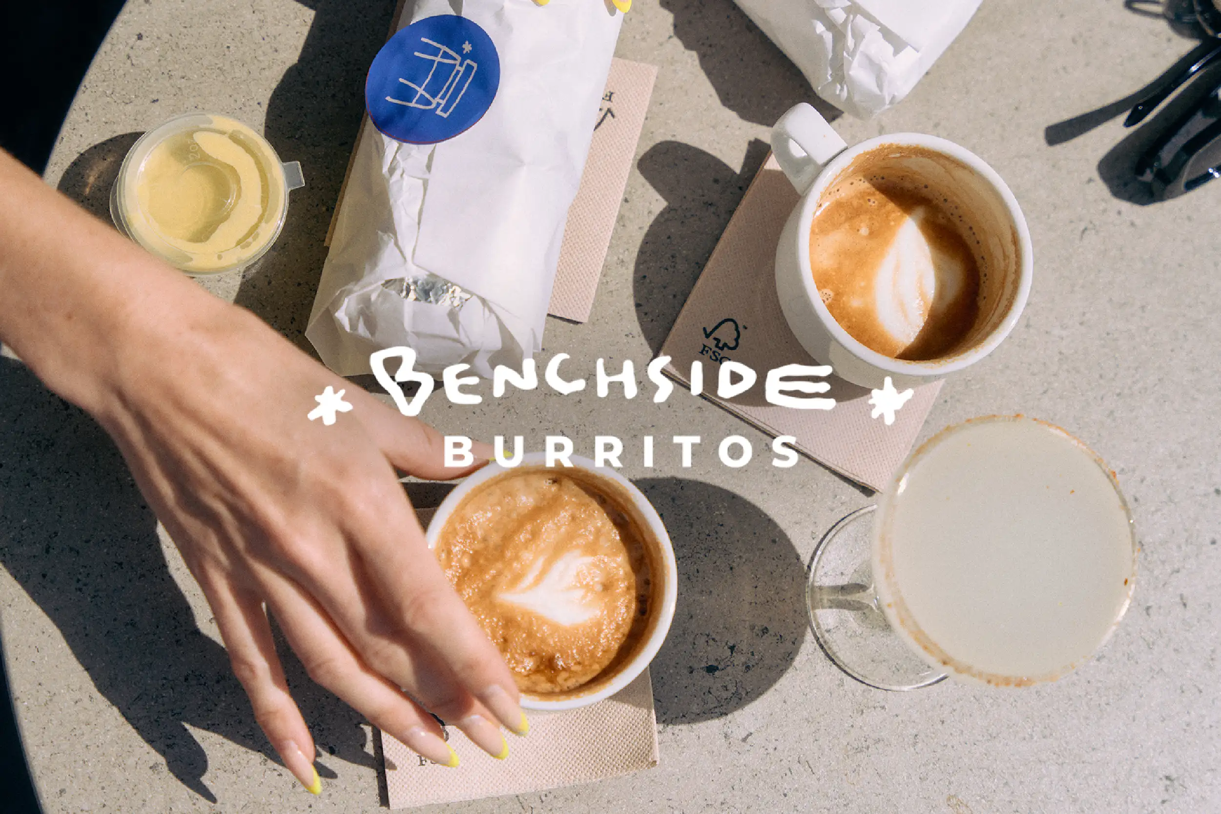 Benchside Burritos wordmark over a woman reaching for a wrapped breakfast burrito with sauce and margarita on a table.