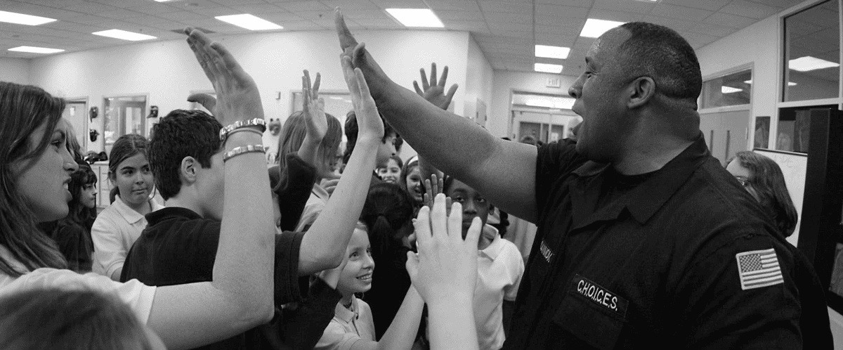 A black and white photo of A black and white photo of Elite founder, Carl Cannon, smiling as he high-fives a group of students in a school. 
