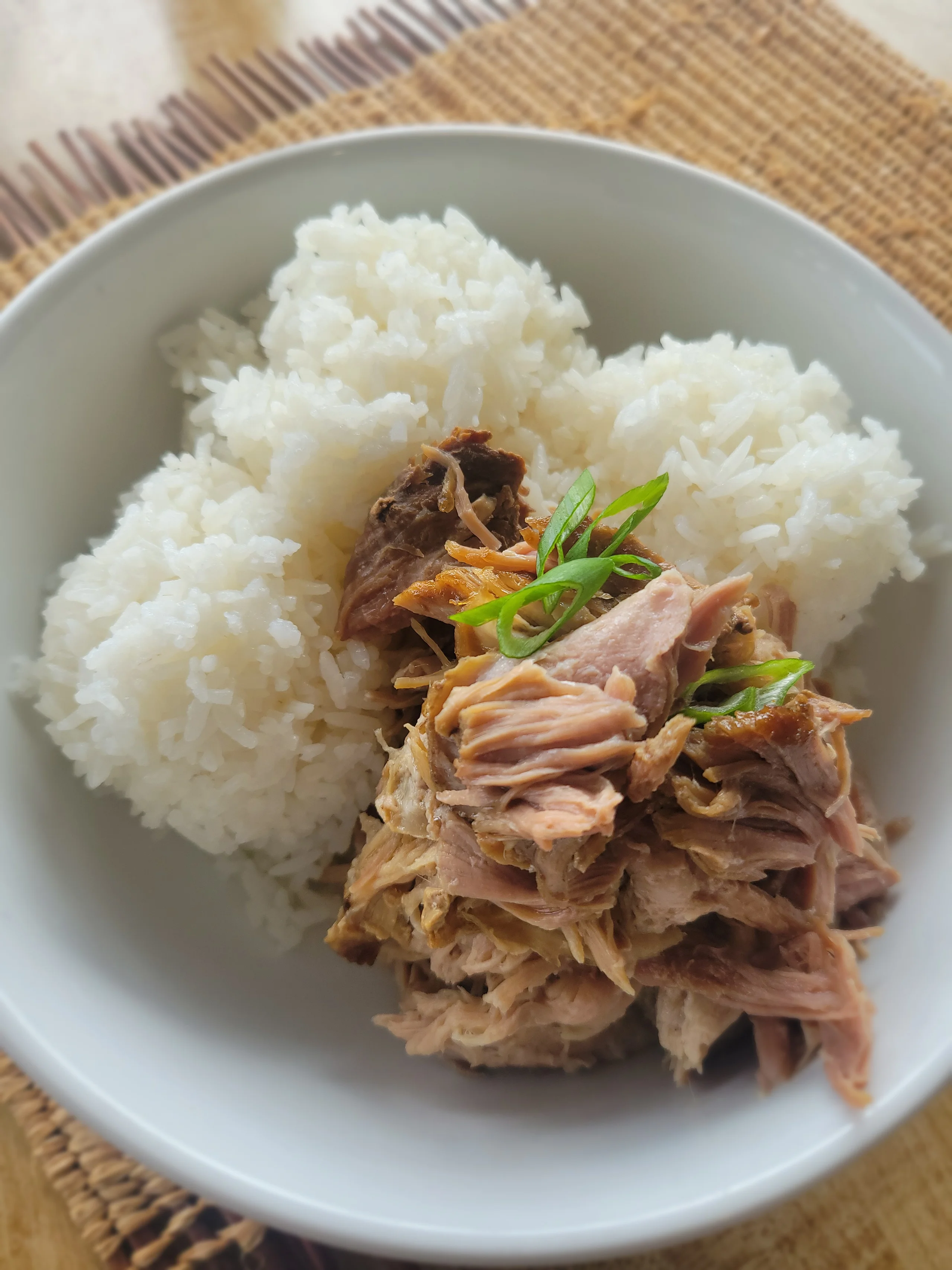A white bowl filled with a large portion of slow-roasted pulled pork garnished with green onions, served next to three scoops of steamed white jasmine rice.